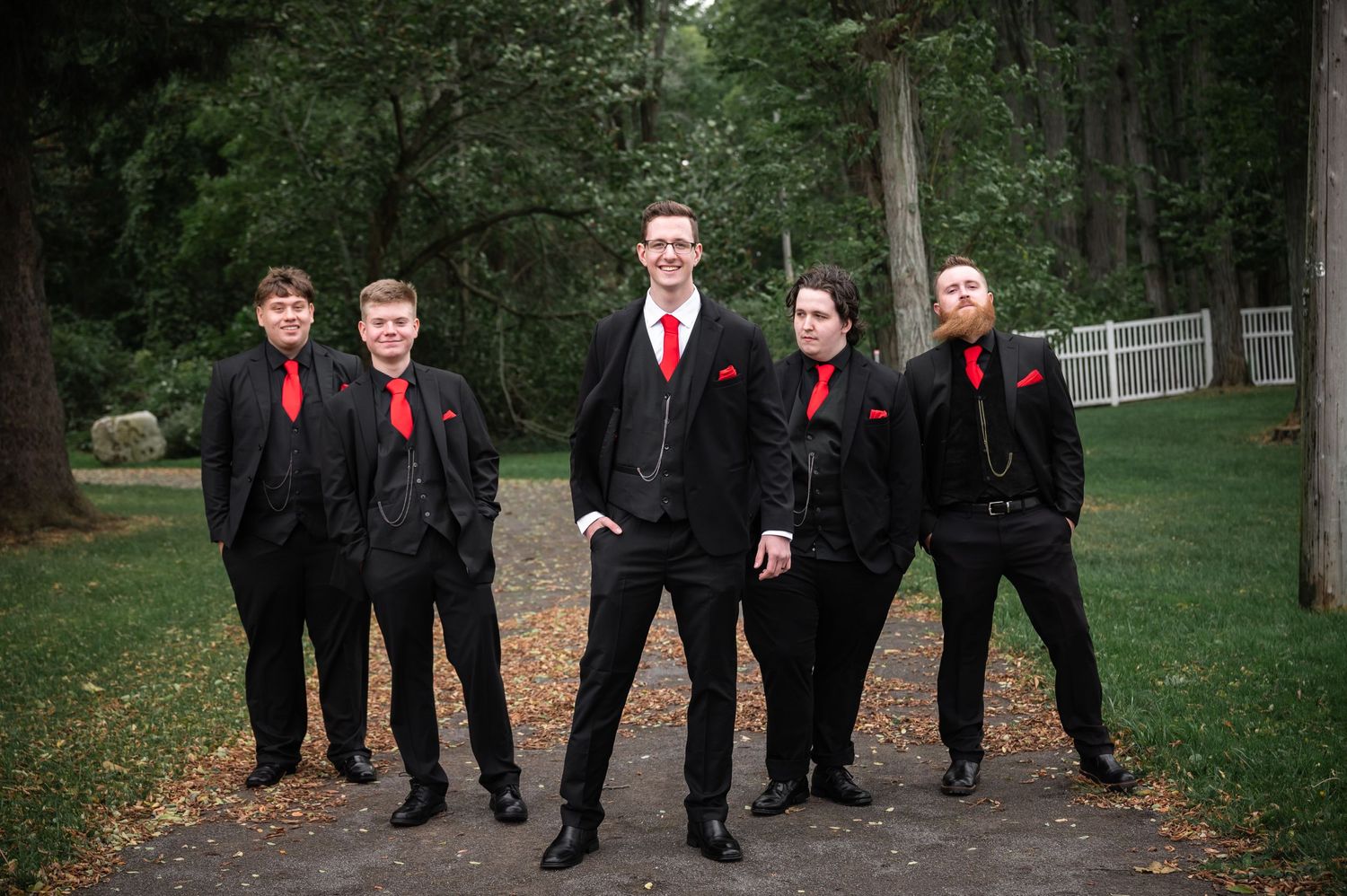 Groomsmen in black suits with red ties pose together on outdoor path.
