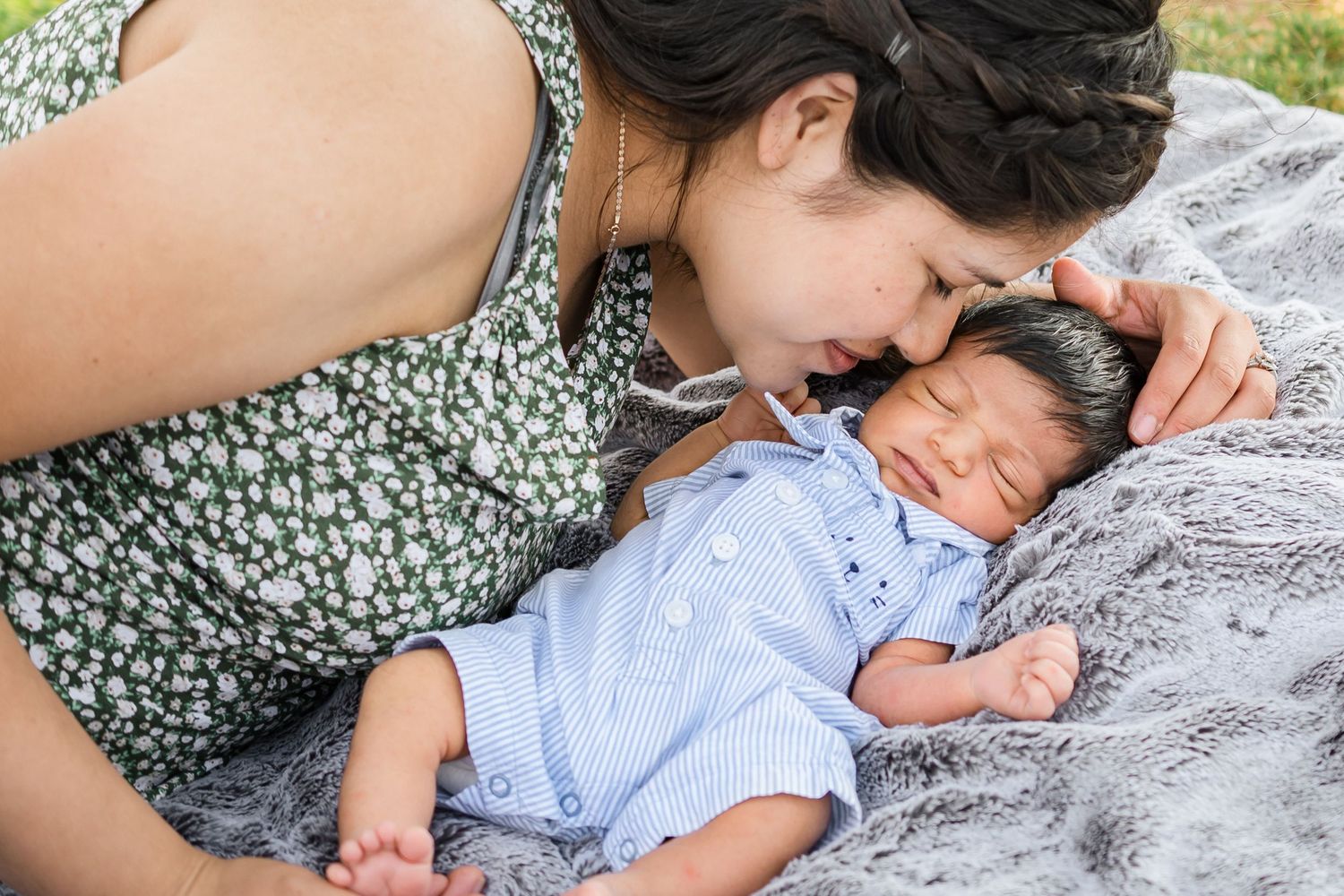 A sleeping baby in a blue outfit lies on a gray blanket while being gently touched by someone in a floral dress.