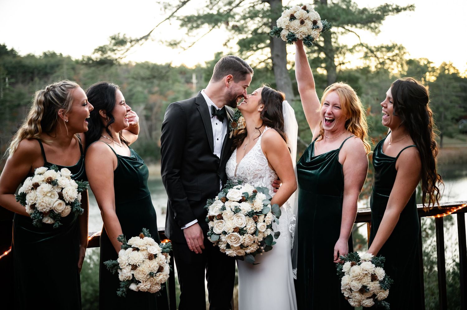 Wedding party shares a joyful moment on a deck railing with white bouquets and forest backdrop at sunset.