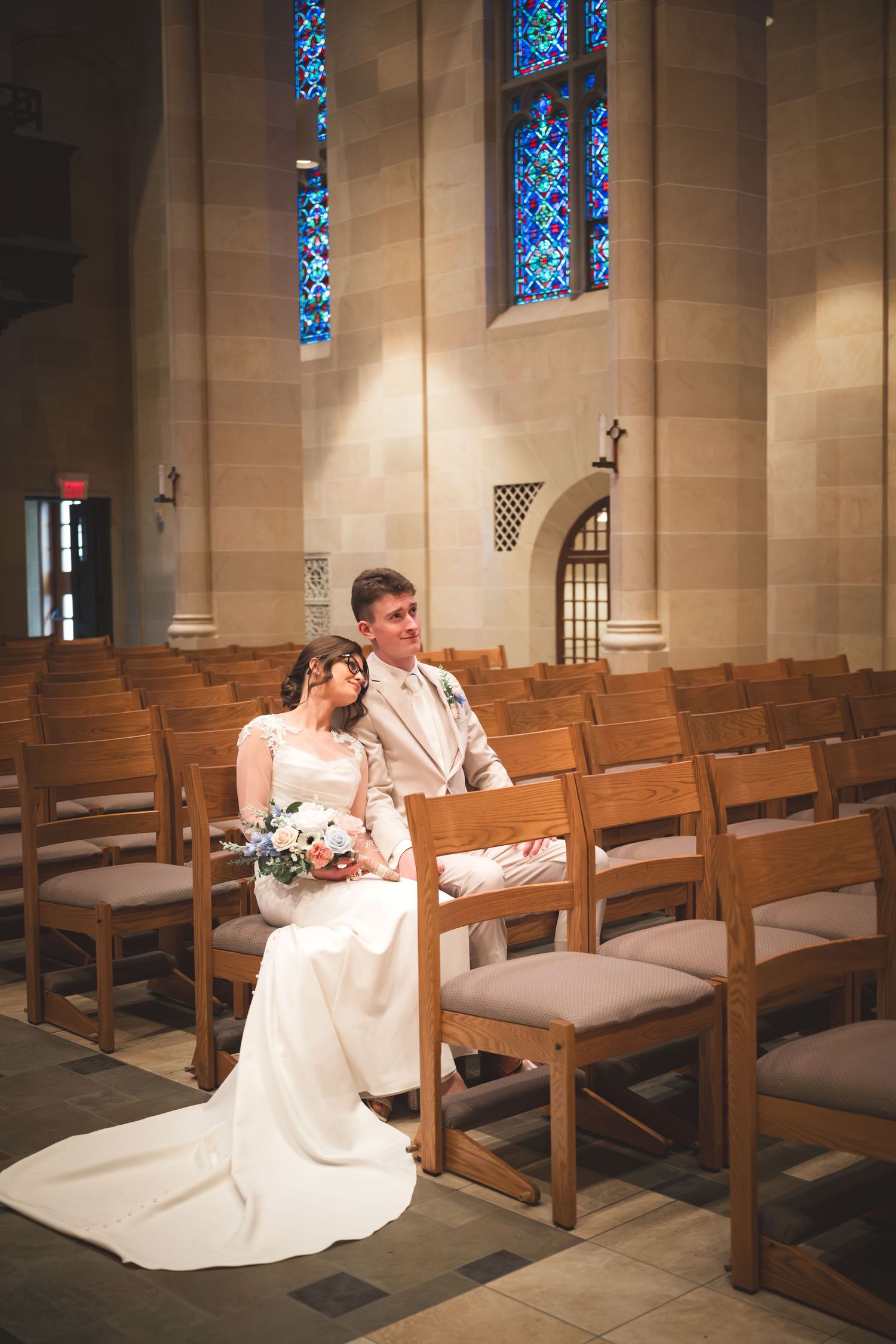 Bride and groom sitting together in church pews, bride holding bouquet, stained glass windows in background.