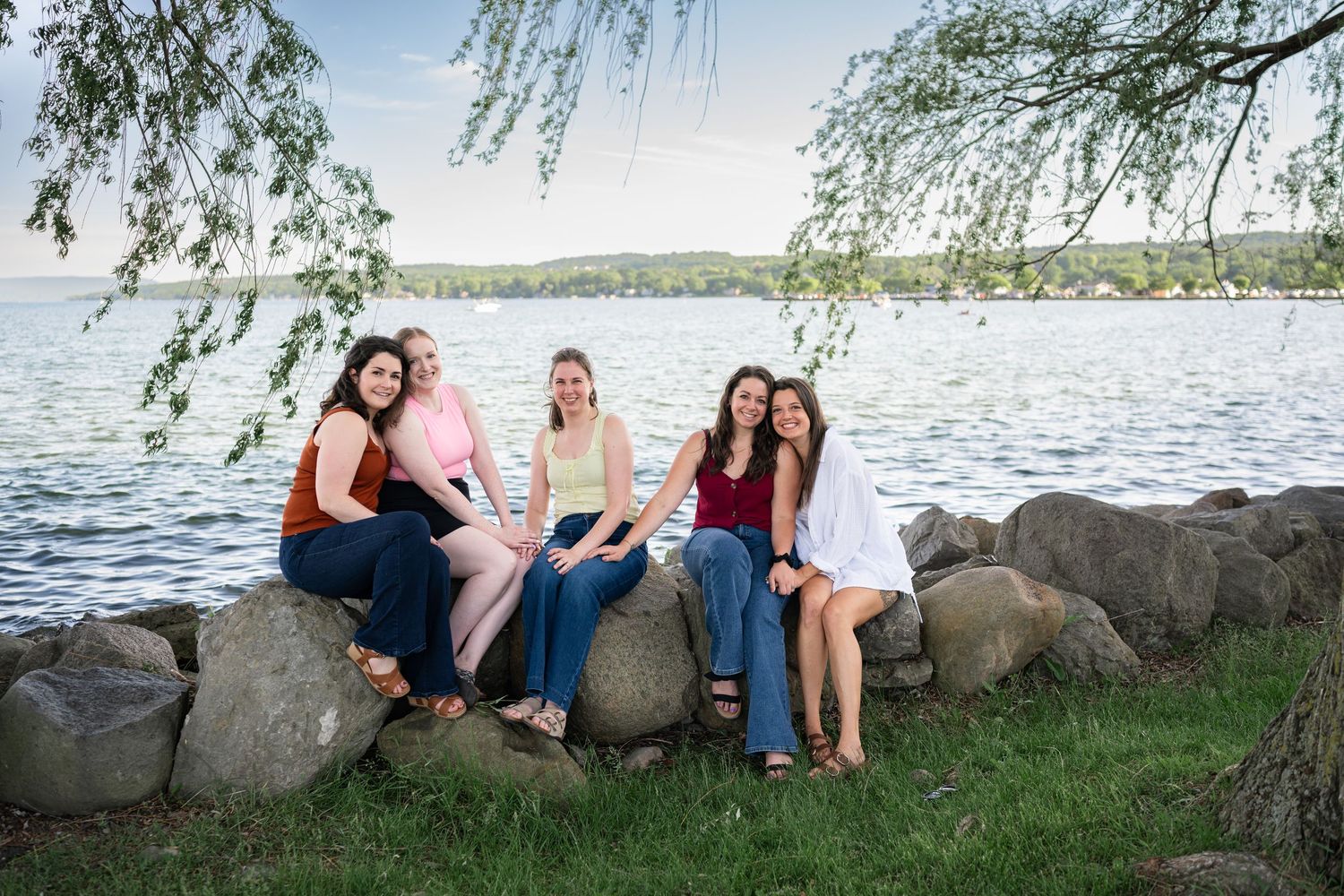 Four young women sitting on rocks by a lake shore, smiling together outdoors on a warm summer day.