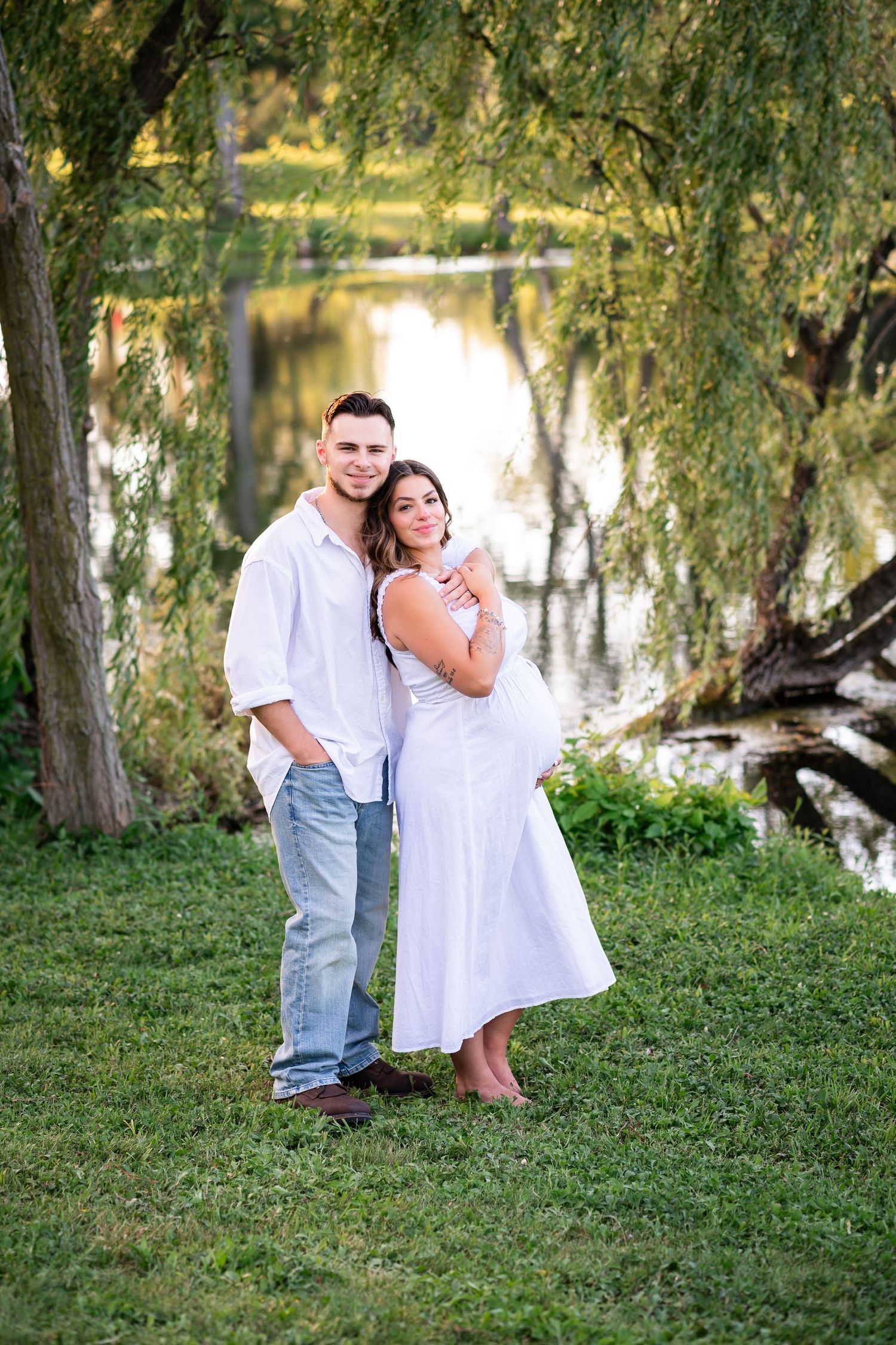 Couple poses by a serene pond under willow trees, woman in white dress with baby bump, man in white shirt and jeans.
