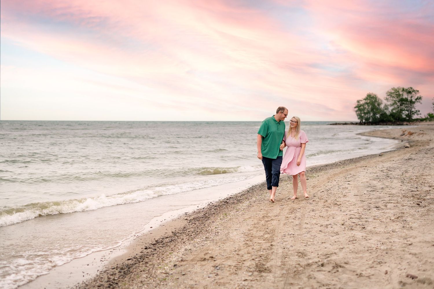 A couple walking along a sandy beach at sunset with pink and purple clouds reflecting on the water.