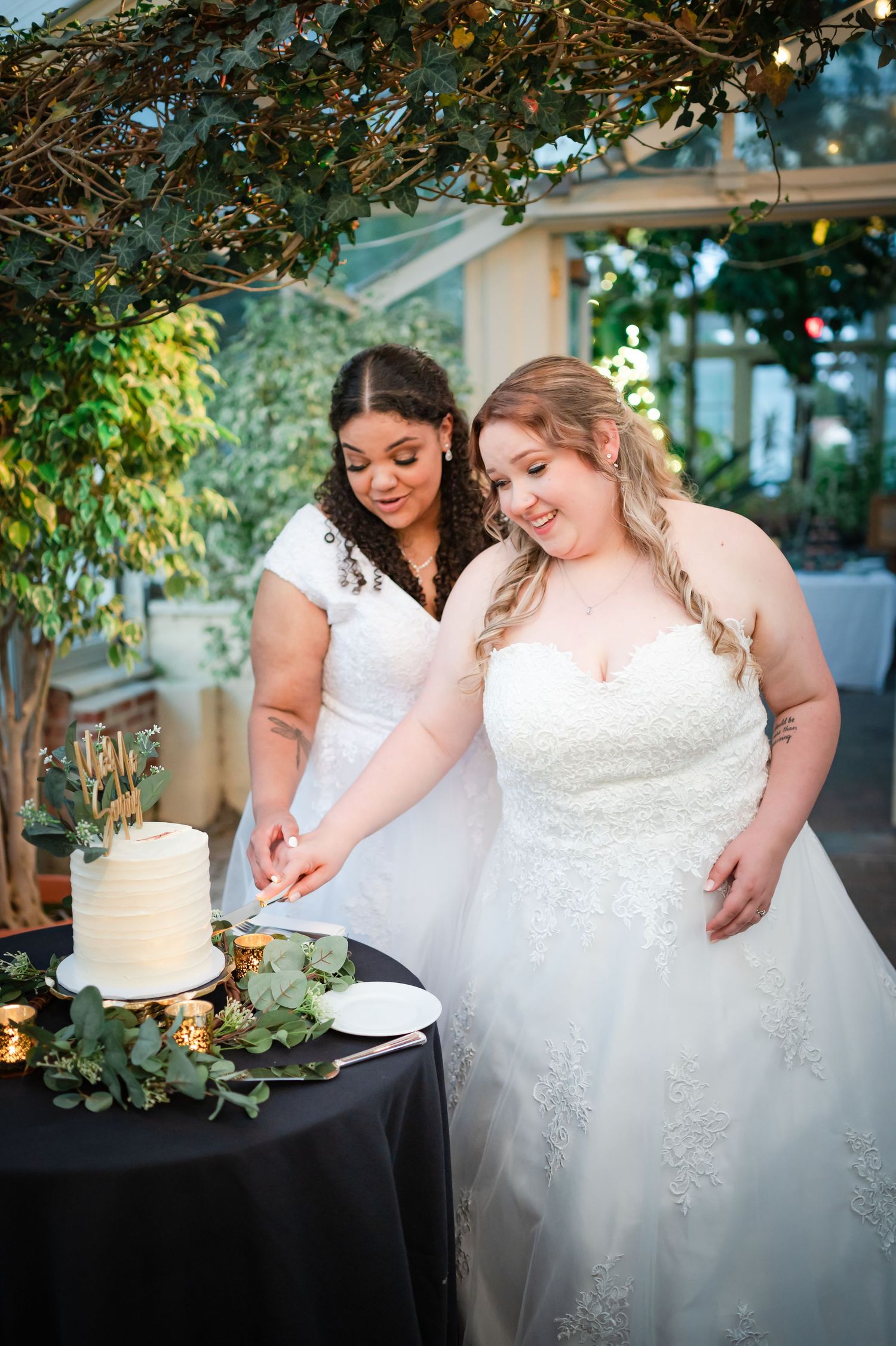 Two brides in white dresses cut their wedding cake together at an outdoor venue with decorative greenery.