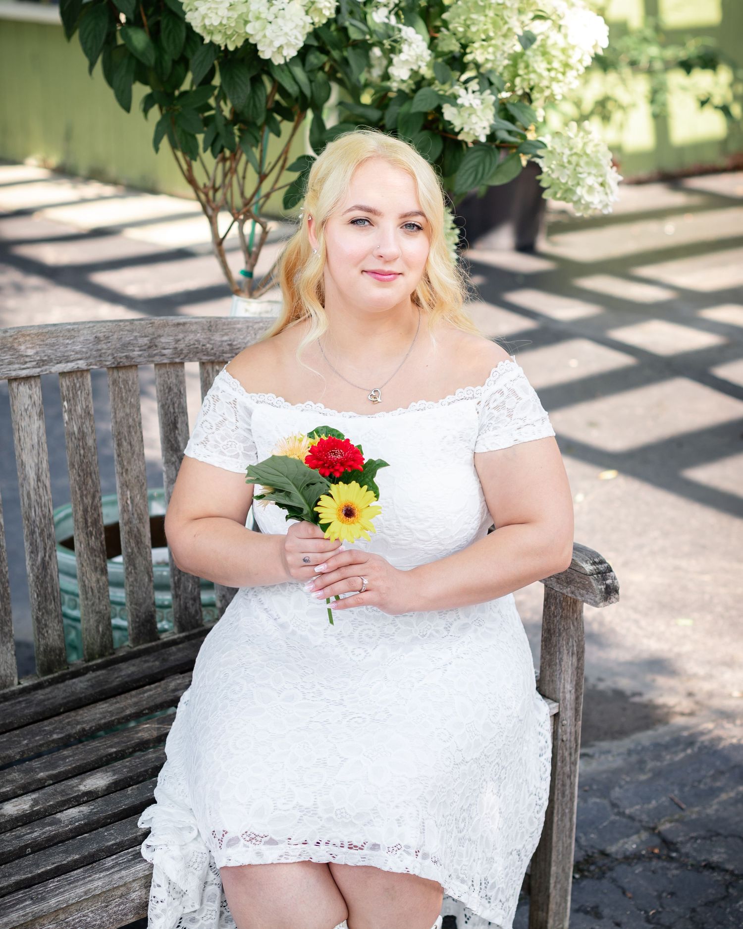 A bride sits on a wooden bench holding colorful flowers beneath white hydrangea blooms.