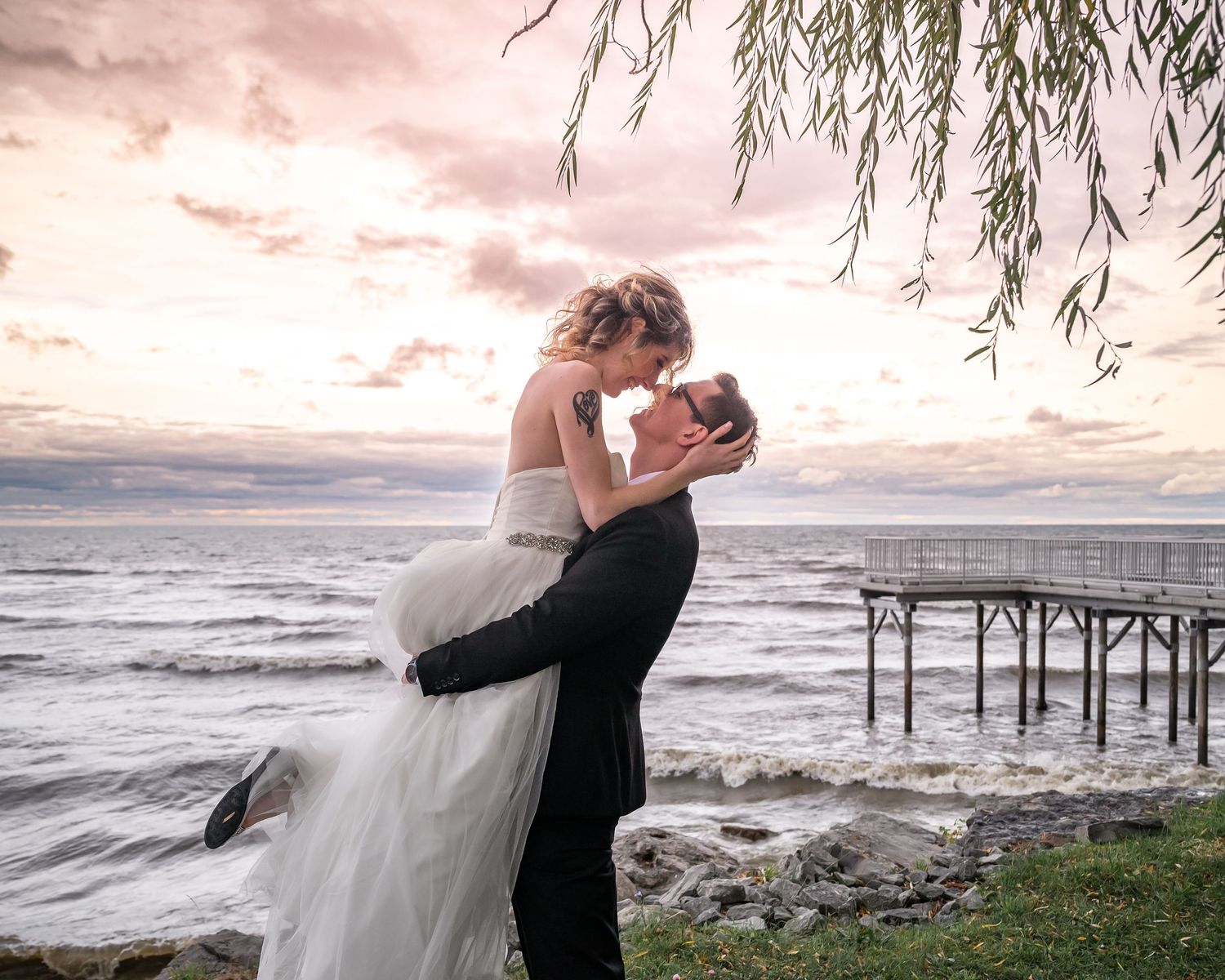 Dramatic sunset wedding photo with couple embracing by ocean pier under willow branches.
