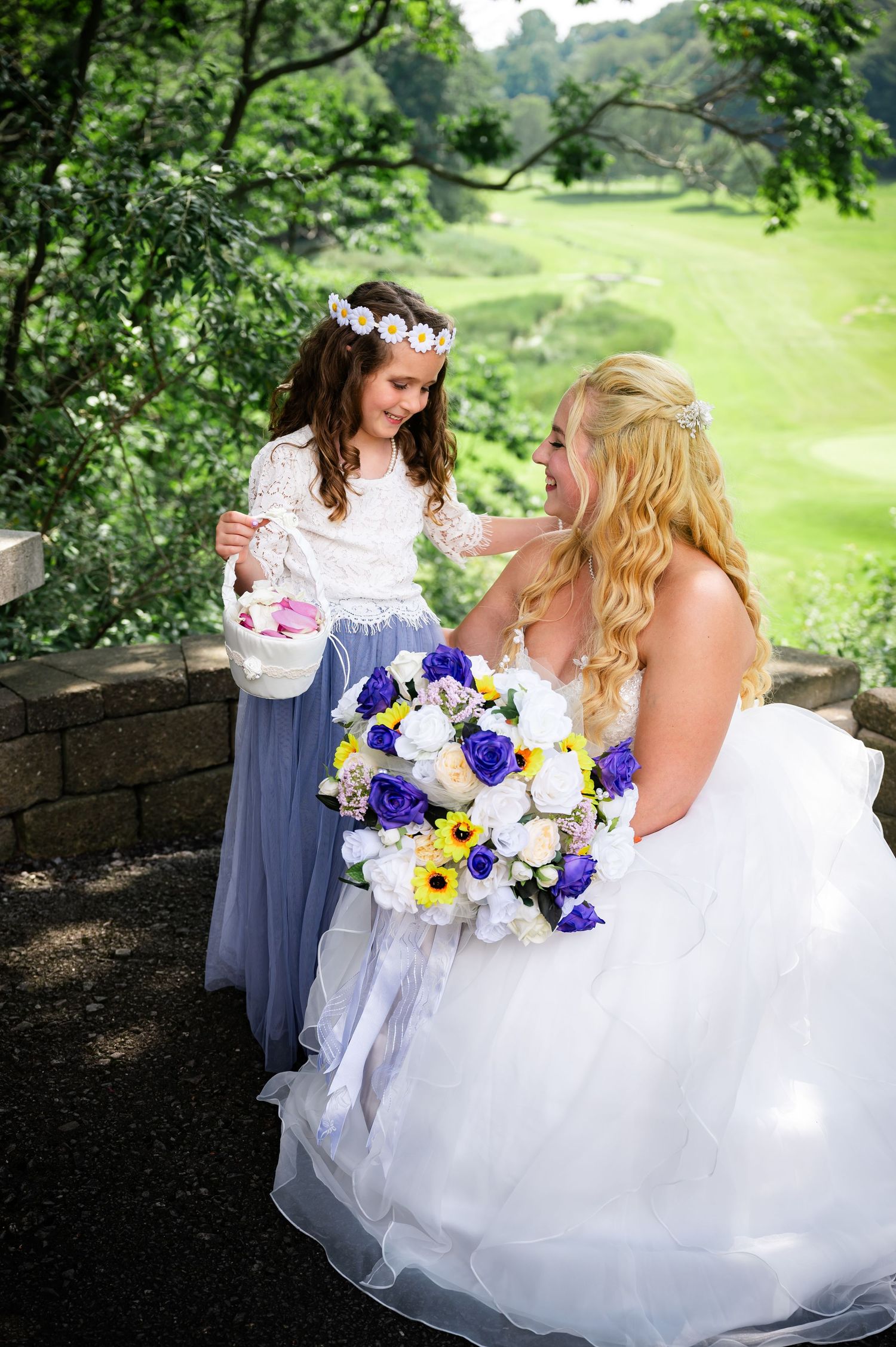 A flower girl in blue and the bride share a sweet moment on stone steps surrounded by greenery.