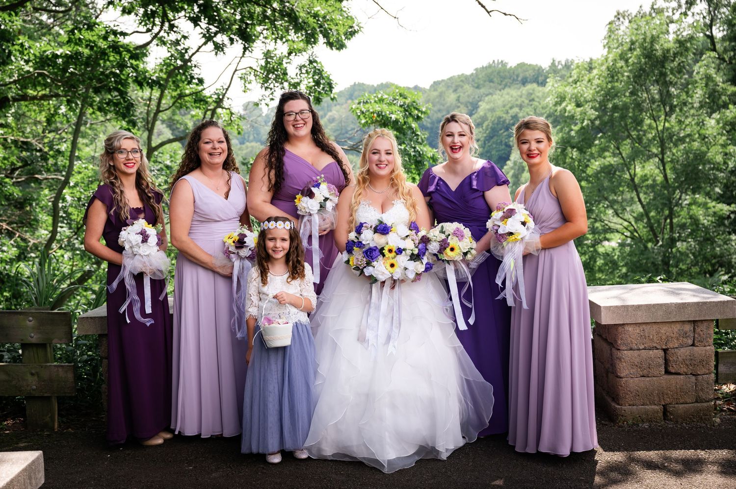Bridal party in purple and lavender dresses poses with flower girls and pet dog on stone overlook.