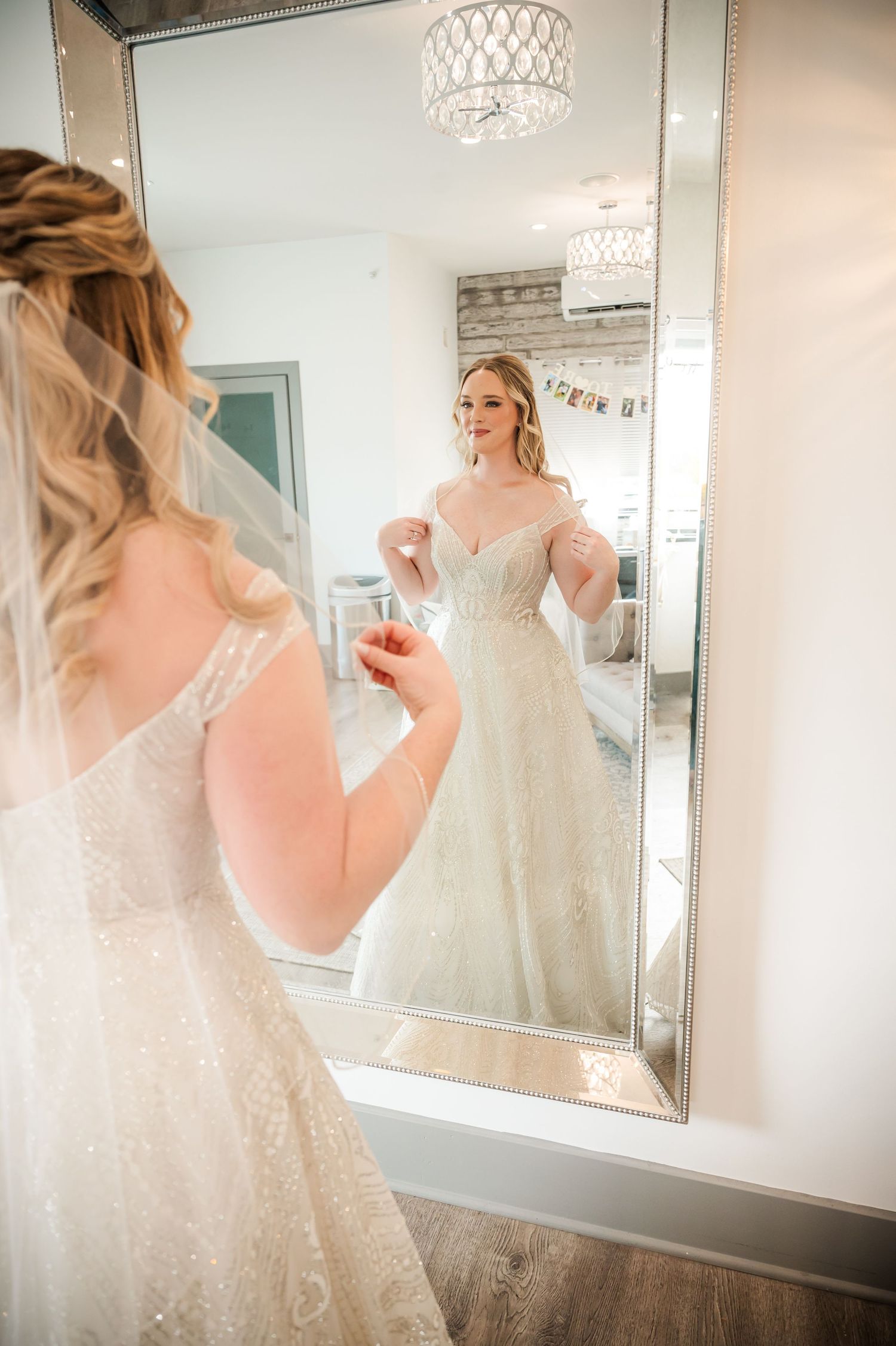 A wedding dress with delicate lace detailing is shown in reflection against elegant mirrors and crystal chandelier lighting.
