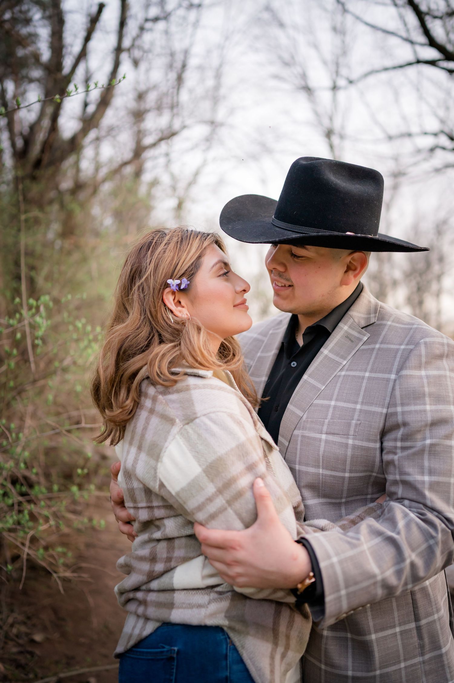 Romantic outdoor couple portrait featuring western style with plaid jacket and black cowboy hat in natural setting.