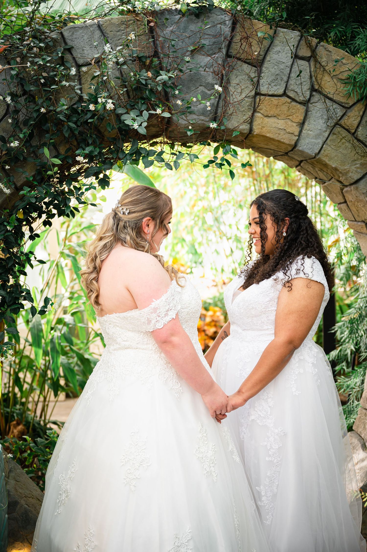 Two brides in white wedding dresses share a tender moment under a stone archway surrounded by lush greenery.