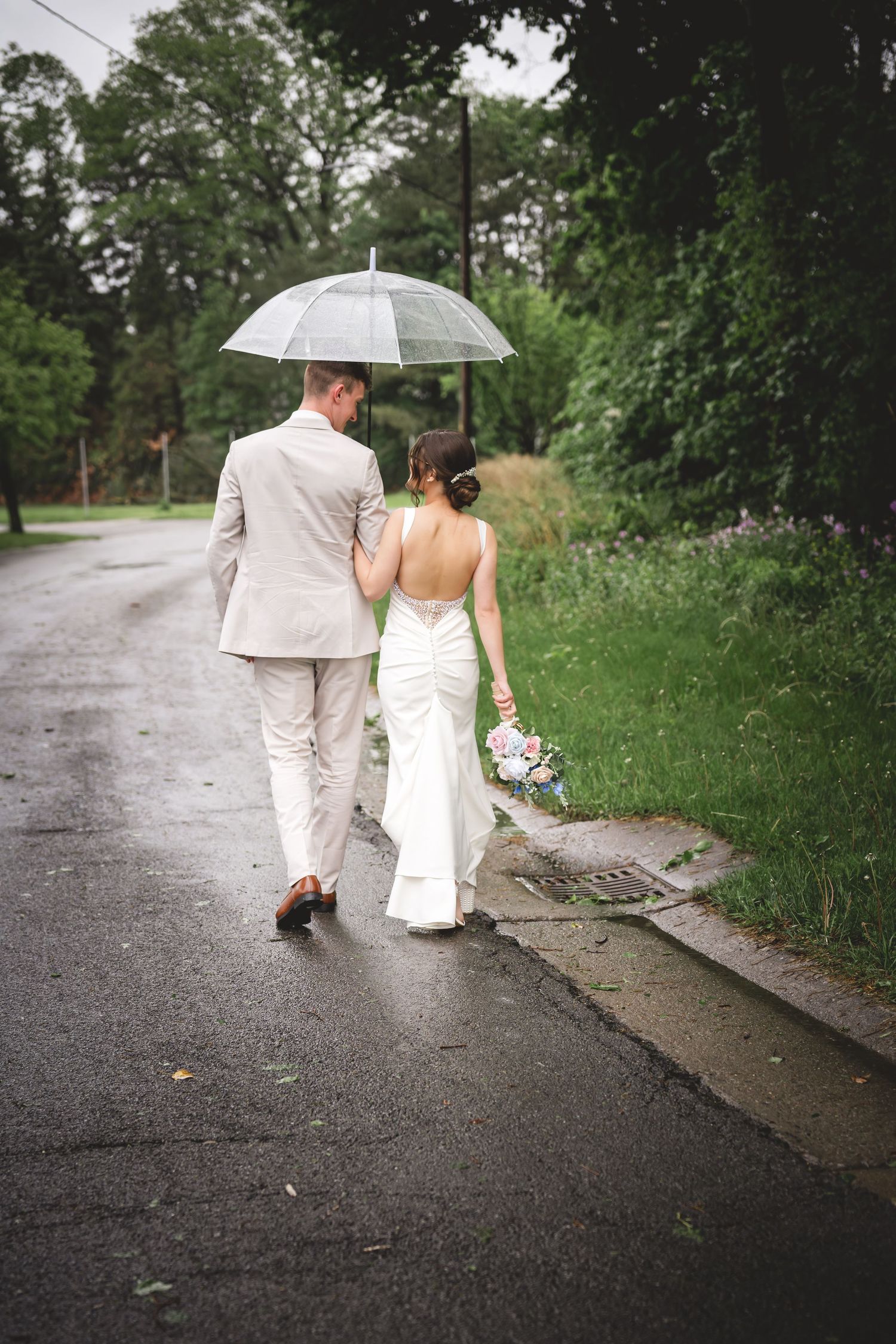 Bride and groom walking under clear umbrella on rainy wedding day, bride in white gown holding bouquet.