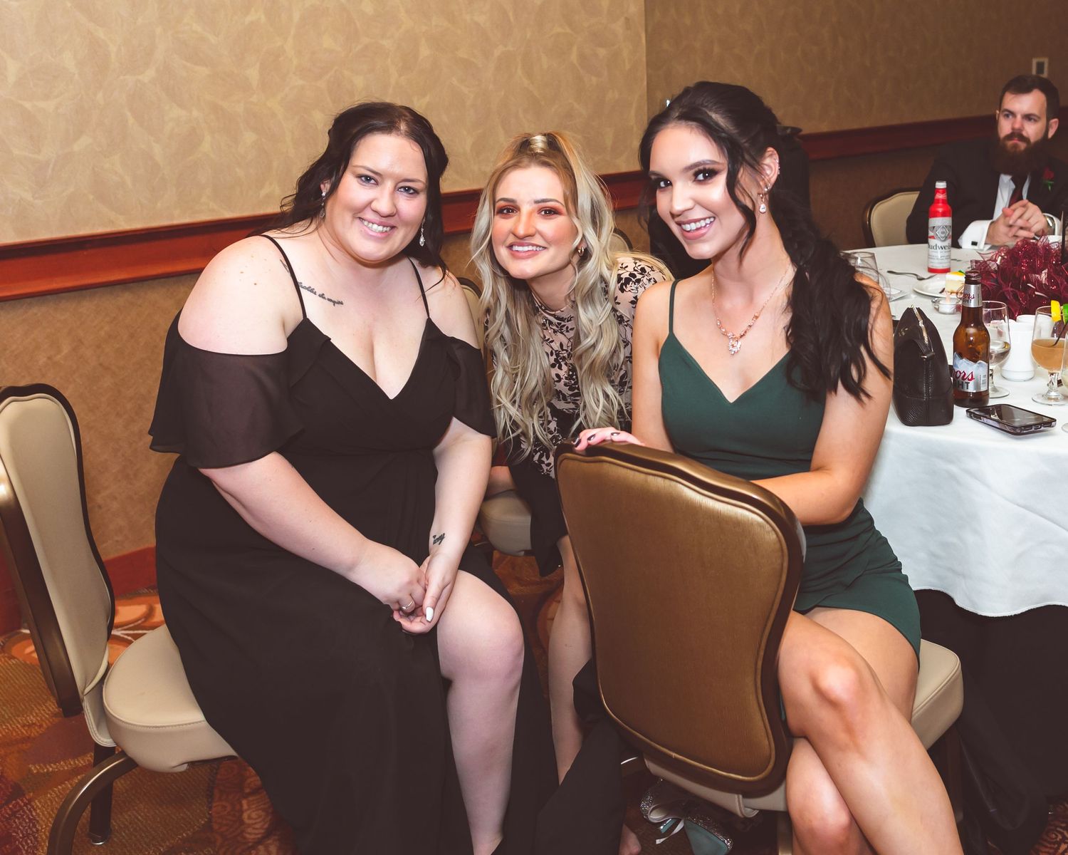 Three wedding guests in formal attire smile while seated at reception table.