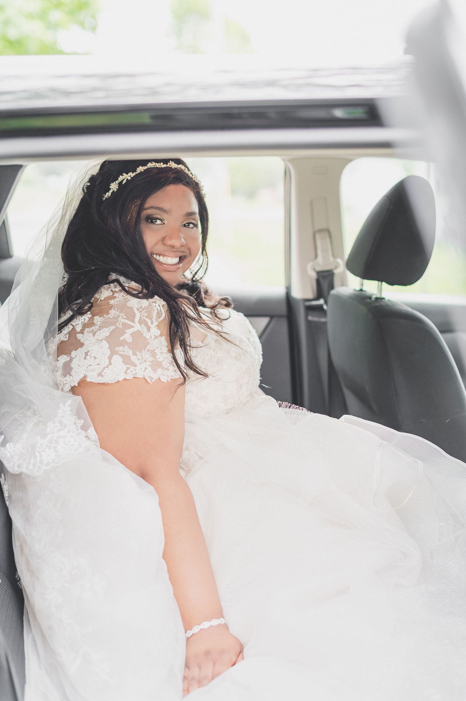 A beaming bride wearing a lace wedding dress sits in the back seat of a car.