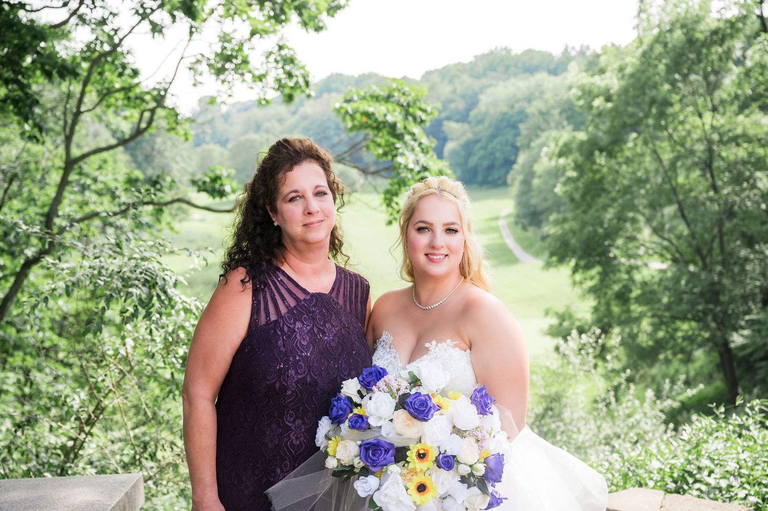 Bride in white gown poses with attendant in purple dress while holding vibrant blue and white bouquet.
