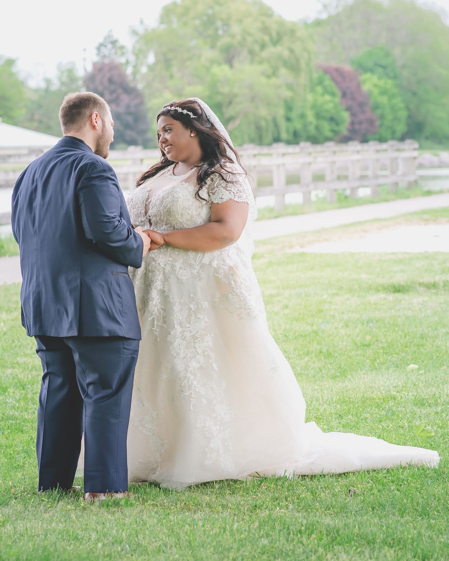 Wedding couple stands together on lawn with wooden fence, bride wearing intricate lace gown with train.