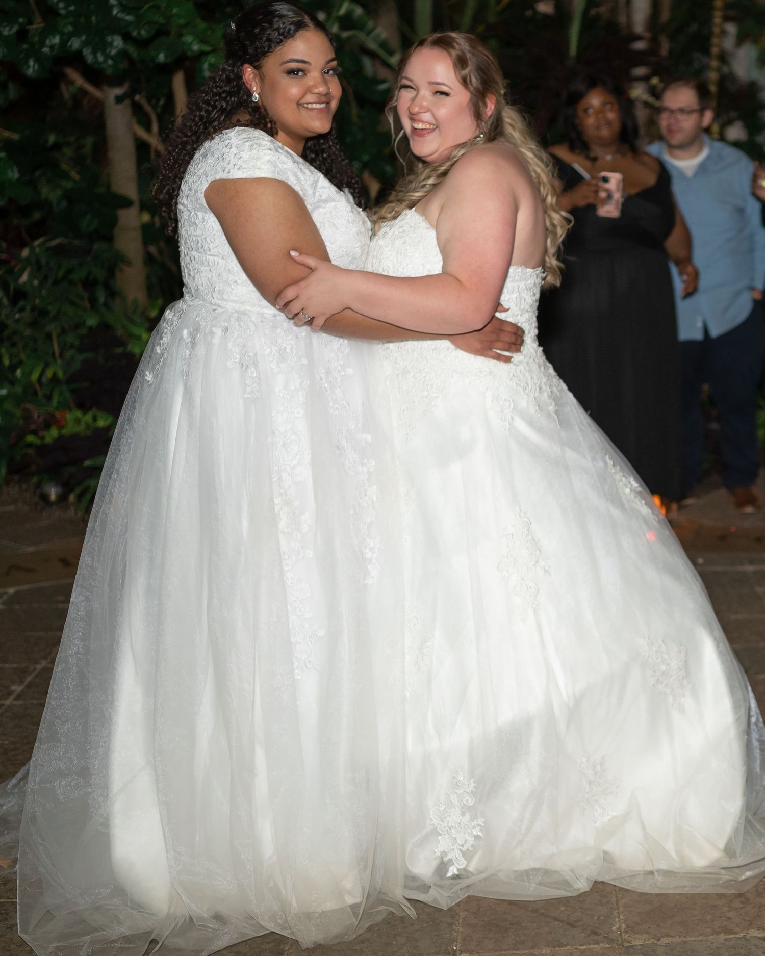 Two brides in matching white wedding gowns embrace on their wedding day.