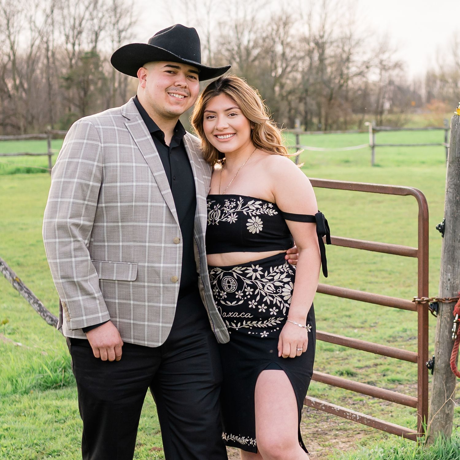 Country-styled couple wearing cowboy hat and plaid blazer embrace outdoors by rustic fence.