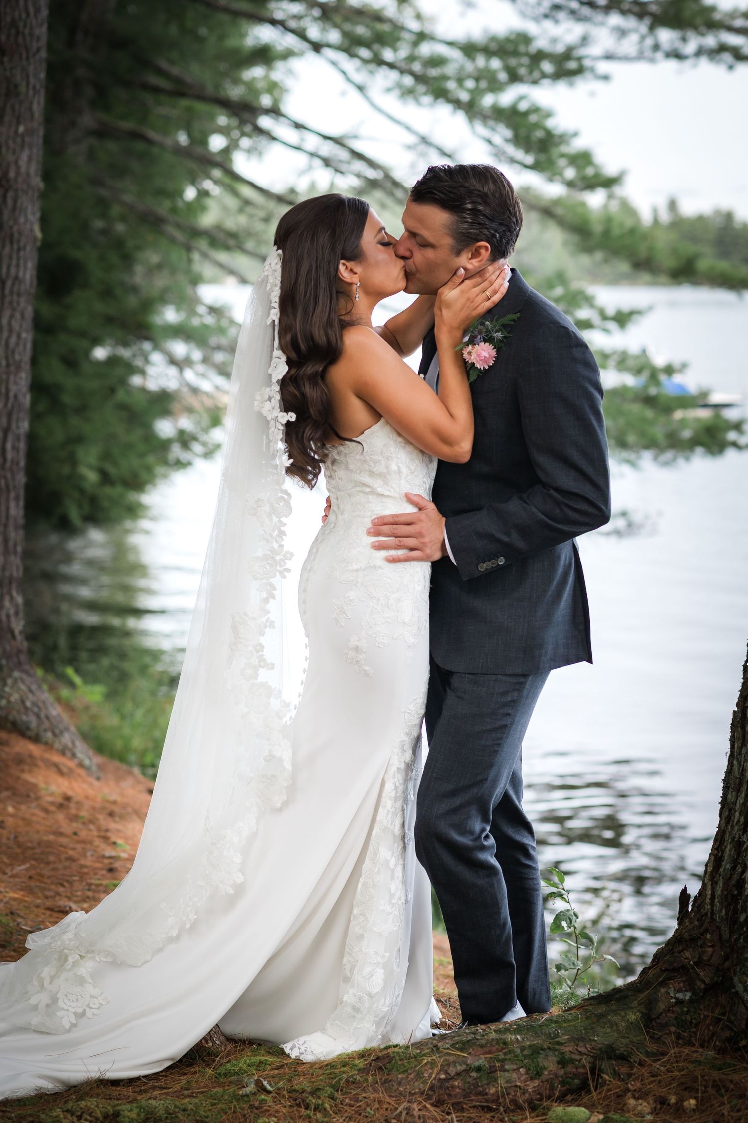 A romantic wedding couple embraces by a serene lake surrounded by pine trees.