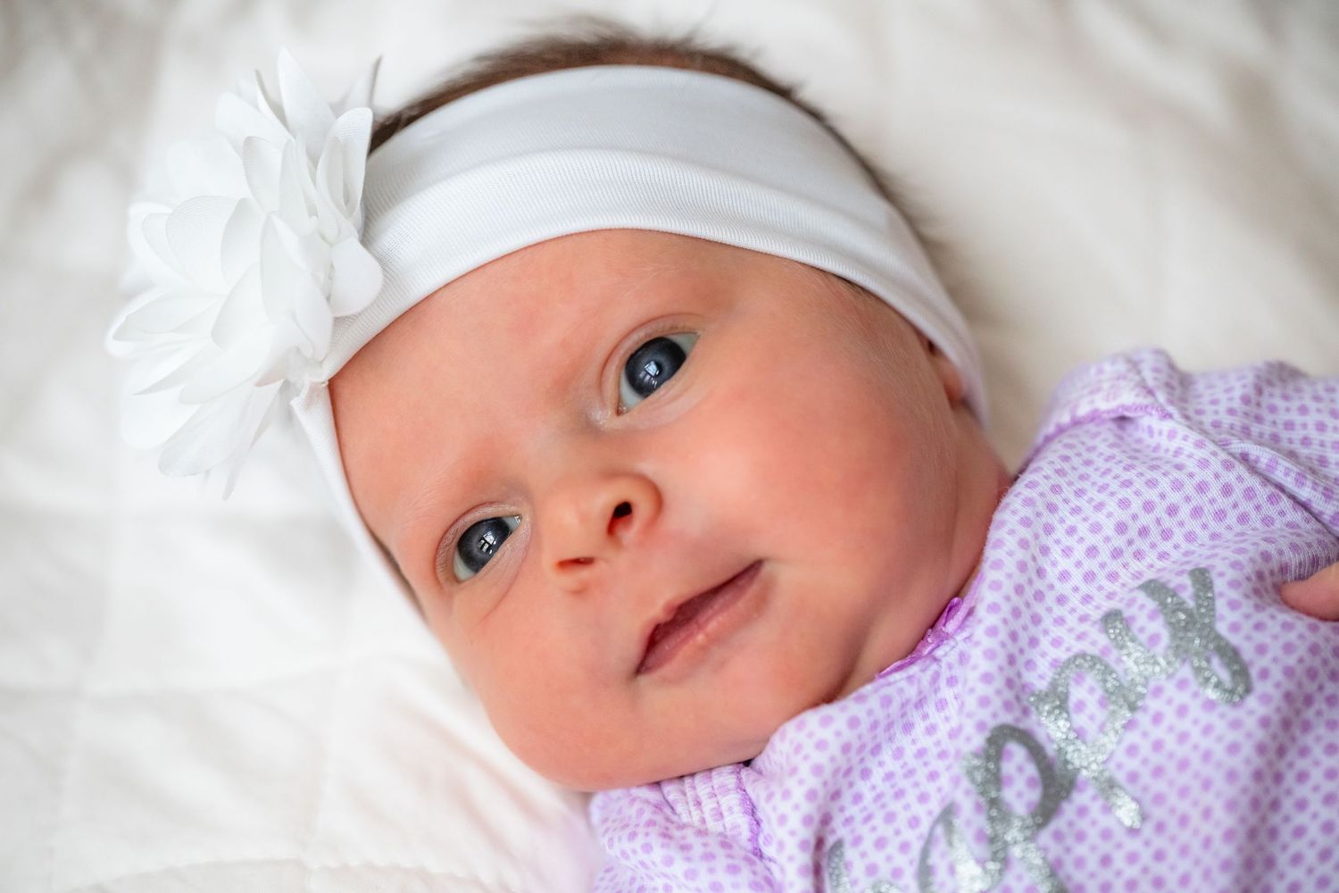 Newborn baby wearing white hat and purple sweater lying on white bedding while looking at camera with bright eyes.
