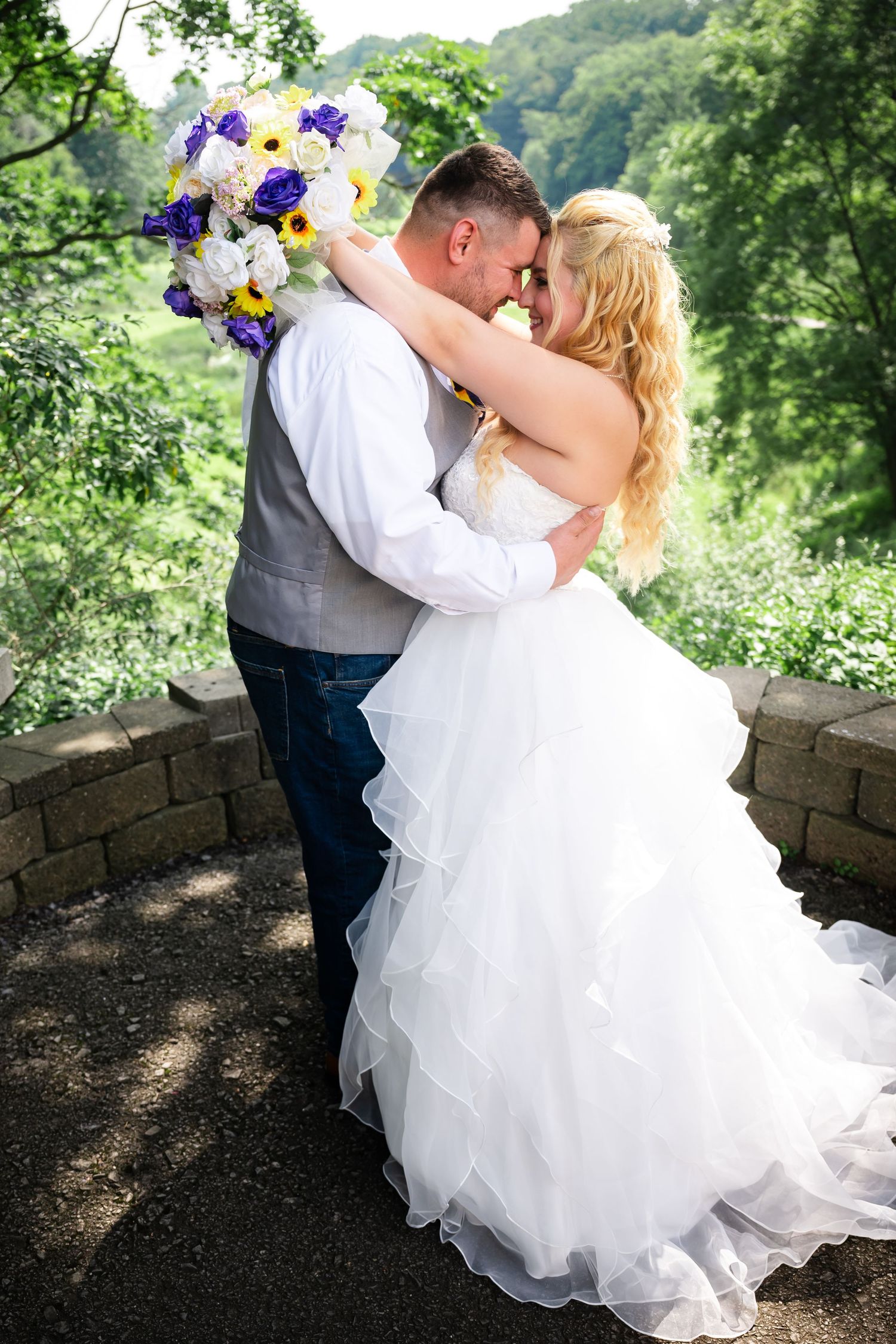 Romantic wedding couple shares tender kiss against stone wall backdrop surrounded by lush greenery and purple bouquet.