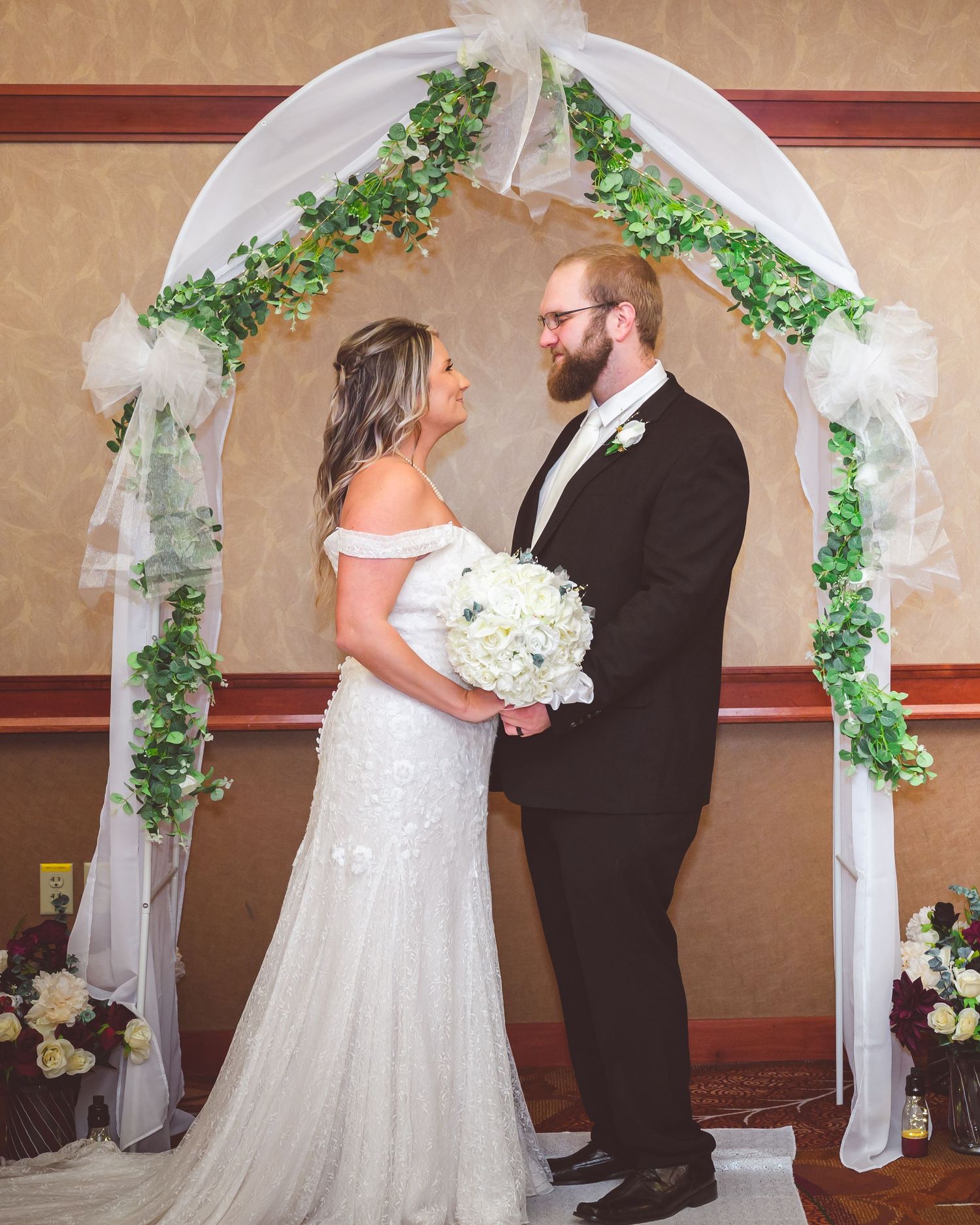 Bride holds white bouquet while posing with groom under decorated wedding arch.