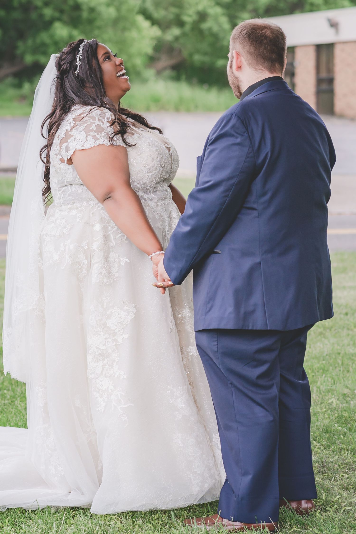 Wedding couple laughs joyfully together on a green lawn, with bride in detailed lace gown and groom in navy suit.