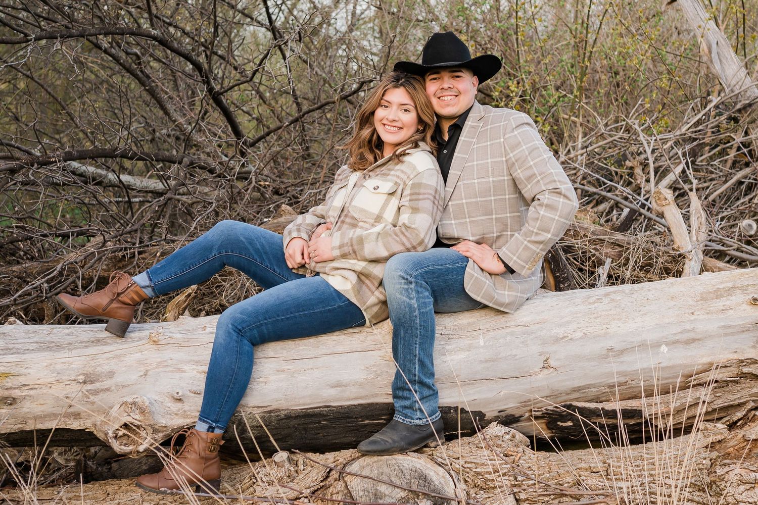 Couple in western wear sits together on fallen log in rustic outdoor setting.