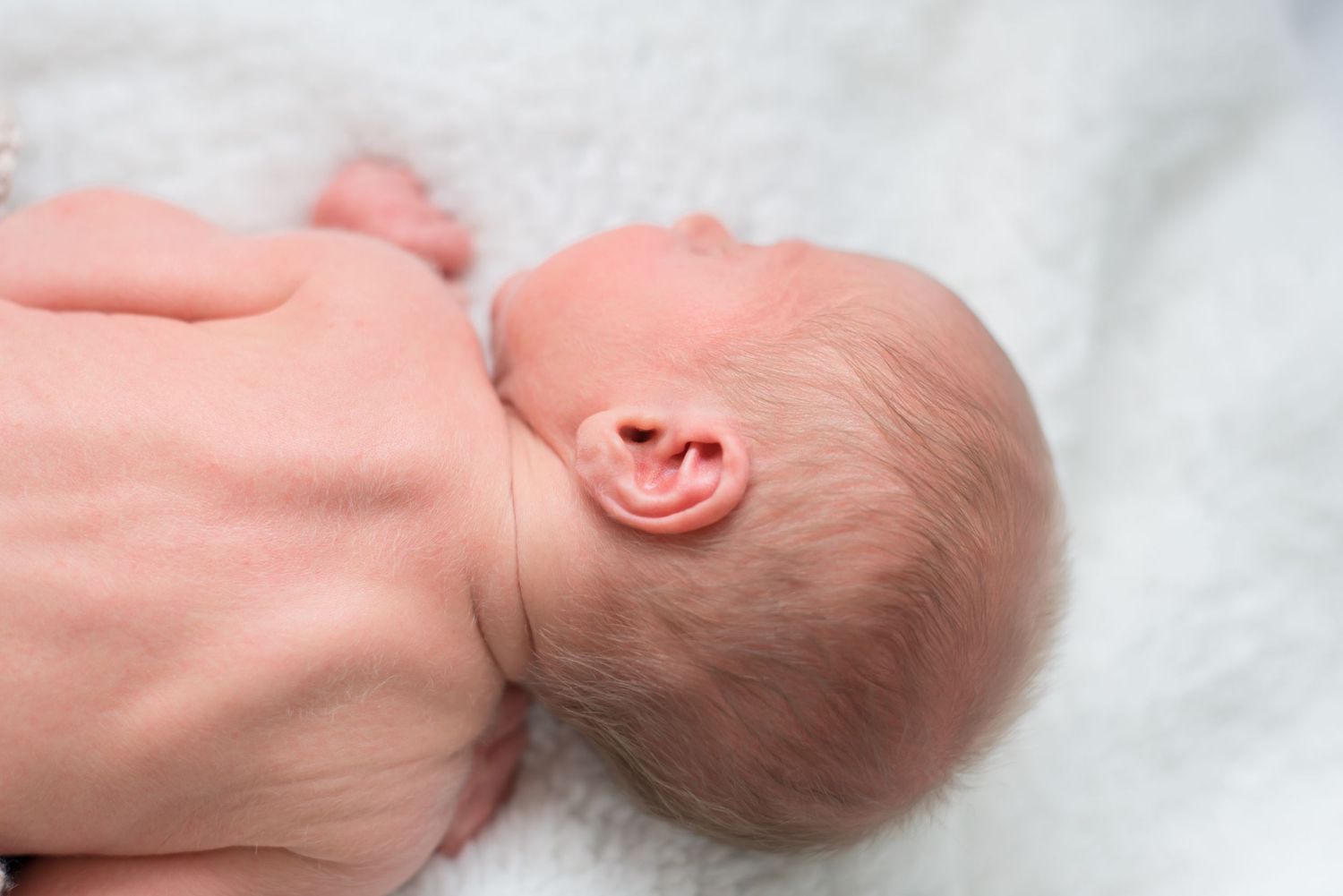 A newborn baby sleeps peacefully on soft white bedding while being gently touched by an adult hand.