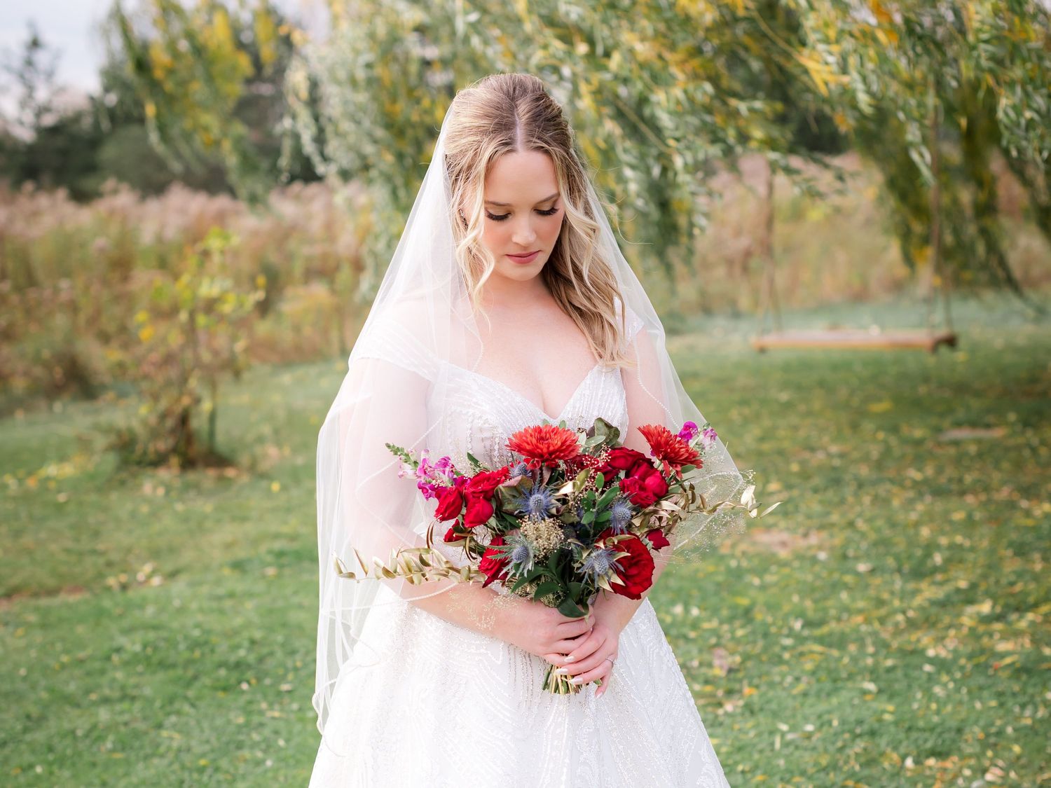 A bride in a flowing white dress and veil holds a vibrant red and burgundy floral bouquet in an outdoor garden setting.
