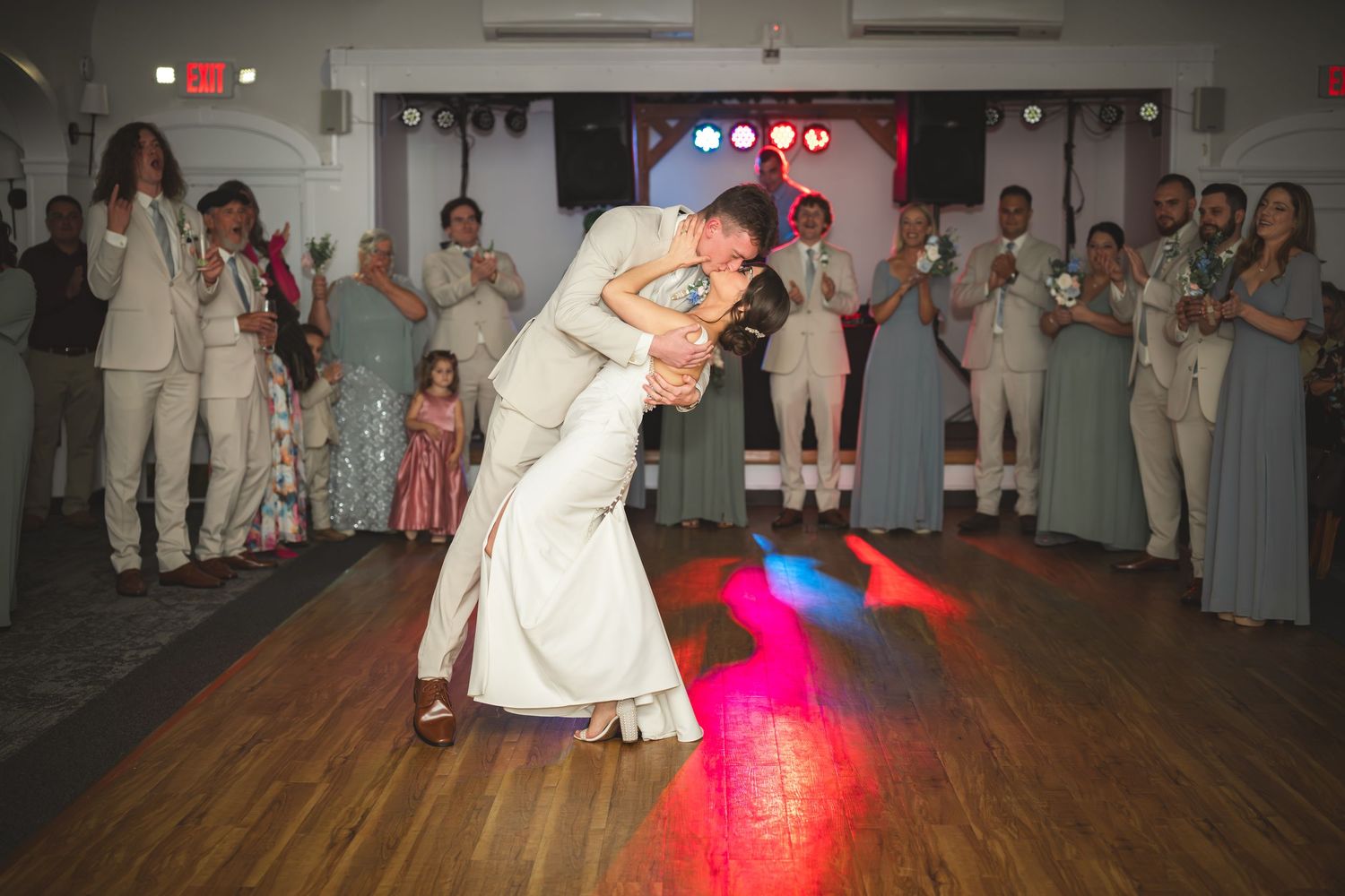 Bride and groom share romantic dip kiss on dance floor surrounded by cheering wedding guests under colorful lights.