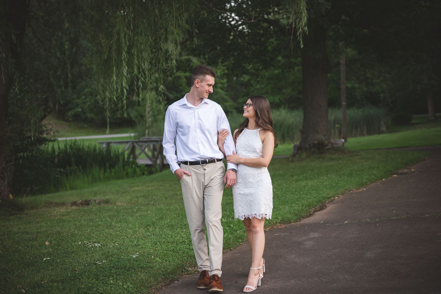 Couple walking hand in hand on a park path, man in white shirt and woman in white dress, surrounded by lush greenery.