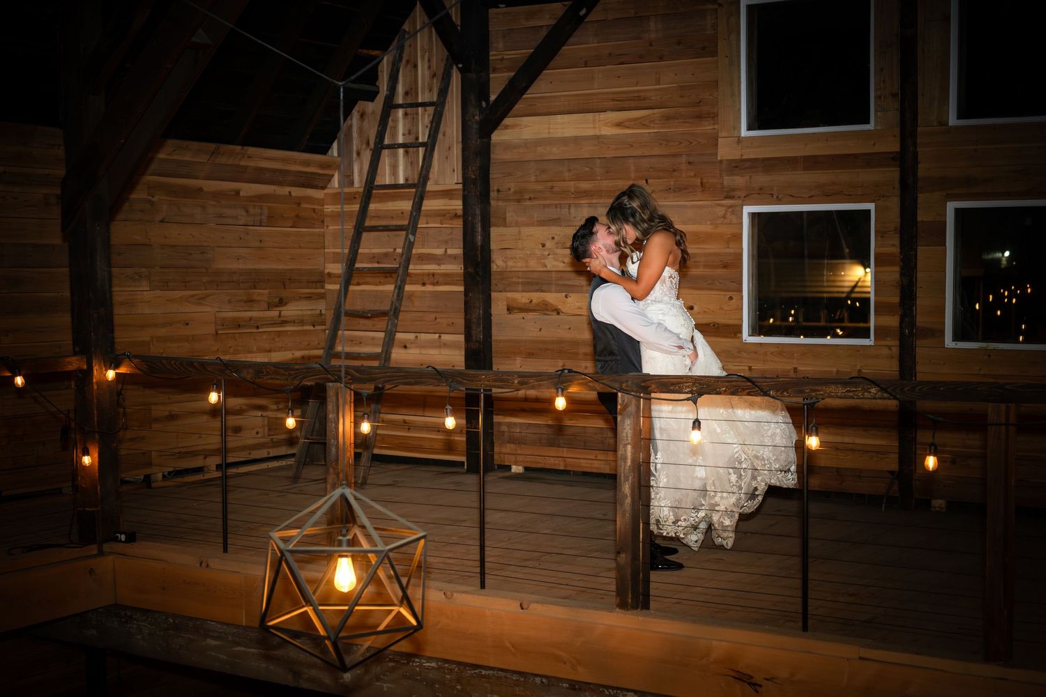 Couple sharing romantic kiss on rustic wooden deck at night, surrounded by warm candlelight and geometric lanterns.