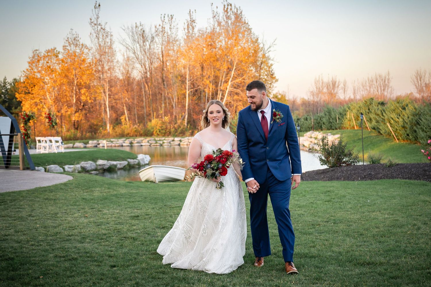 A couple in wedding attire stands on a lawn with autumn trees and a pond in the background during golden hour.