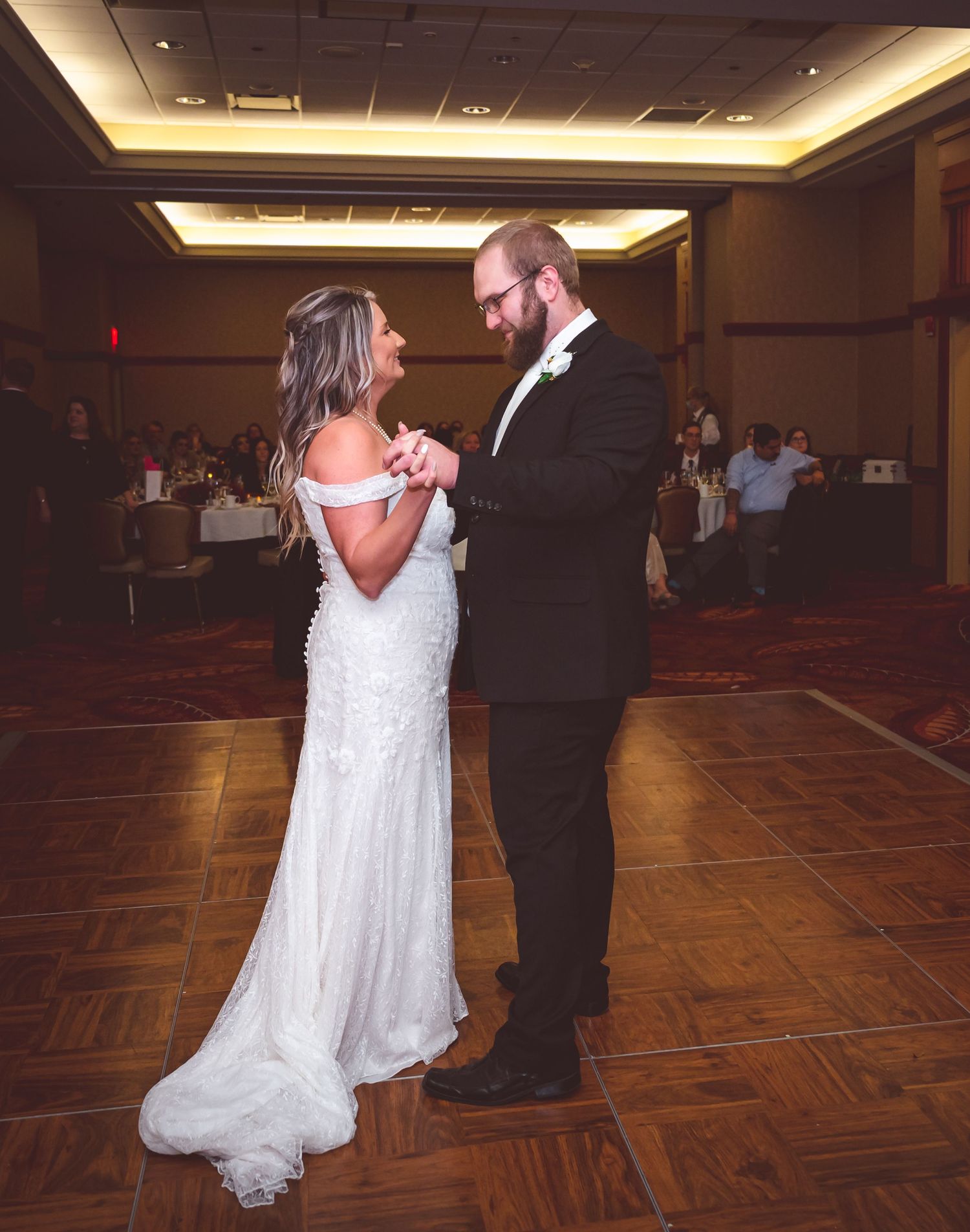 Wedding couple shares first dance on wooden floor in dimly lit reception hall.