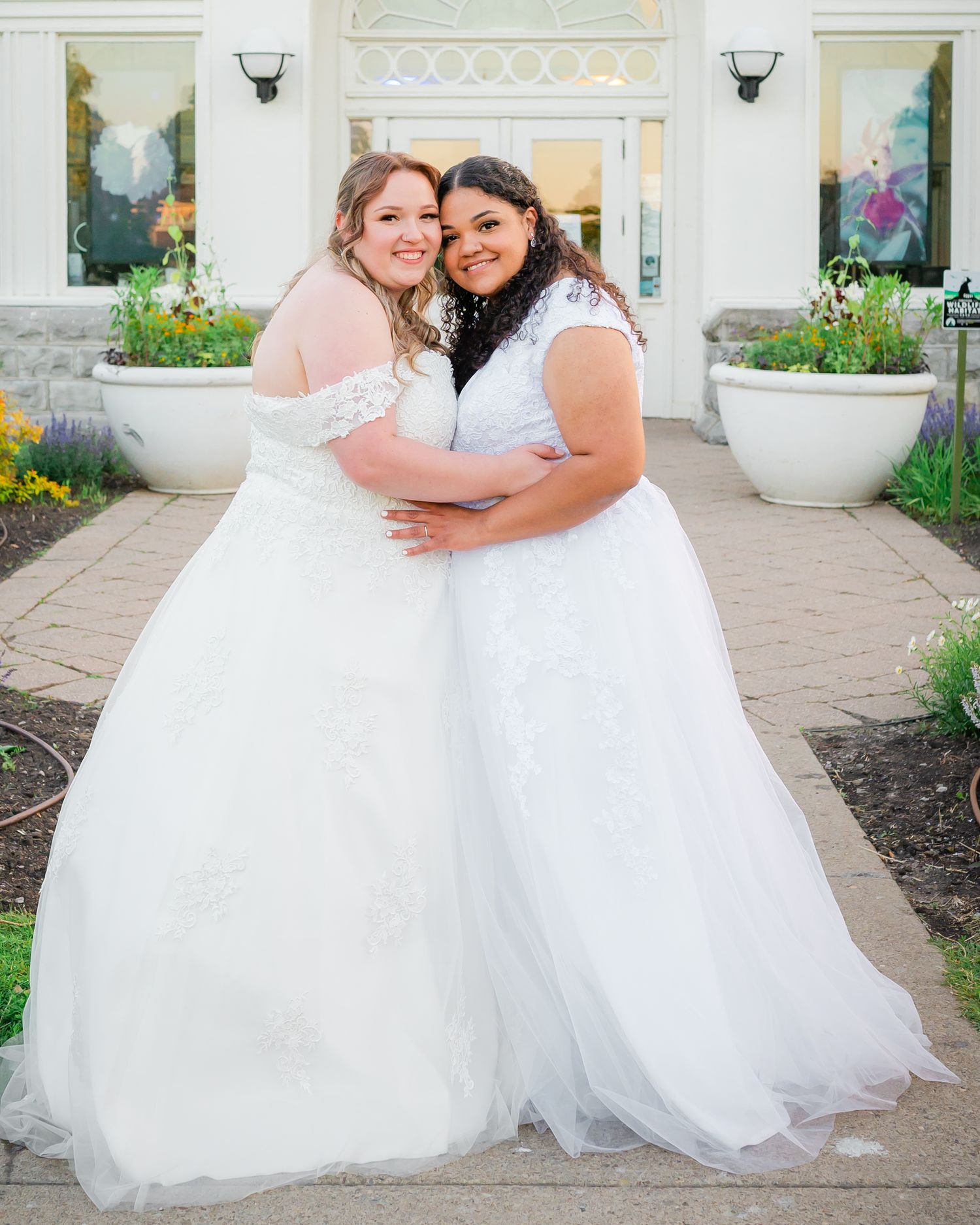 Two brides in white wedding gowns embrace and smile in front of decorative garden planters.