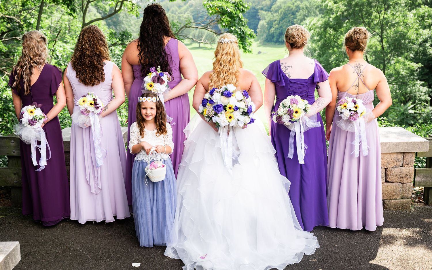 Wedding party in various shades of purple dresses pose with bouquets against a scenic backdrop.