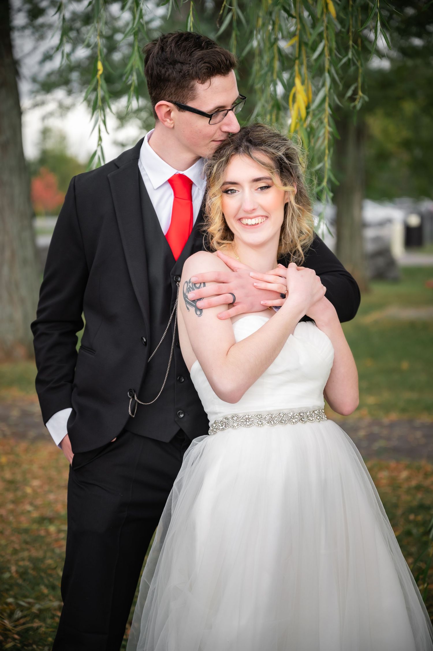 A tender moment between newlyweds under a willow tree, the bride wearing a sparkling belted white gown.