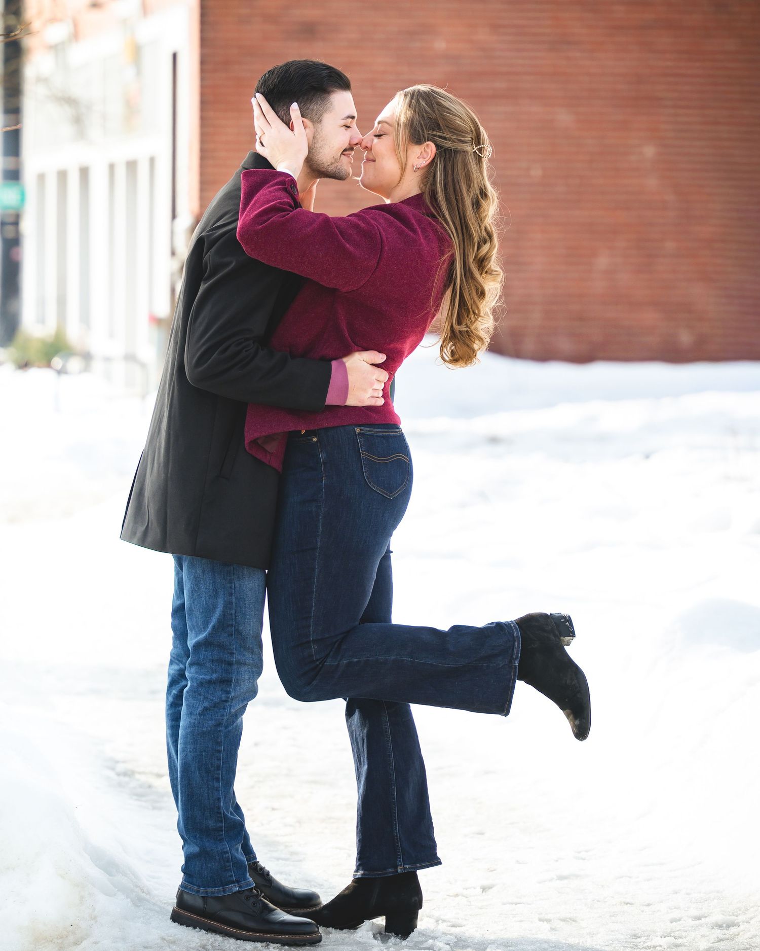 Romantic couple kissing in a snowy outdoor setting, woman lifting her leg in a joyful embrace.