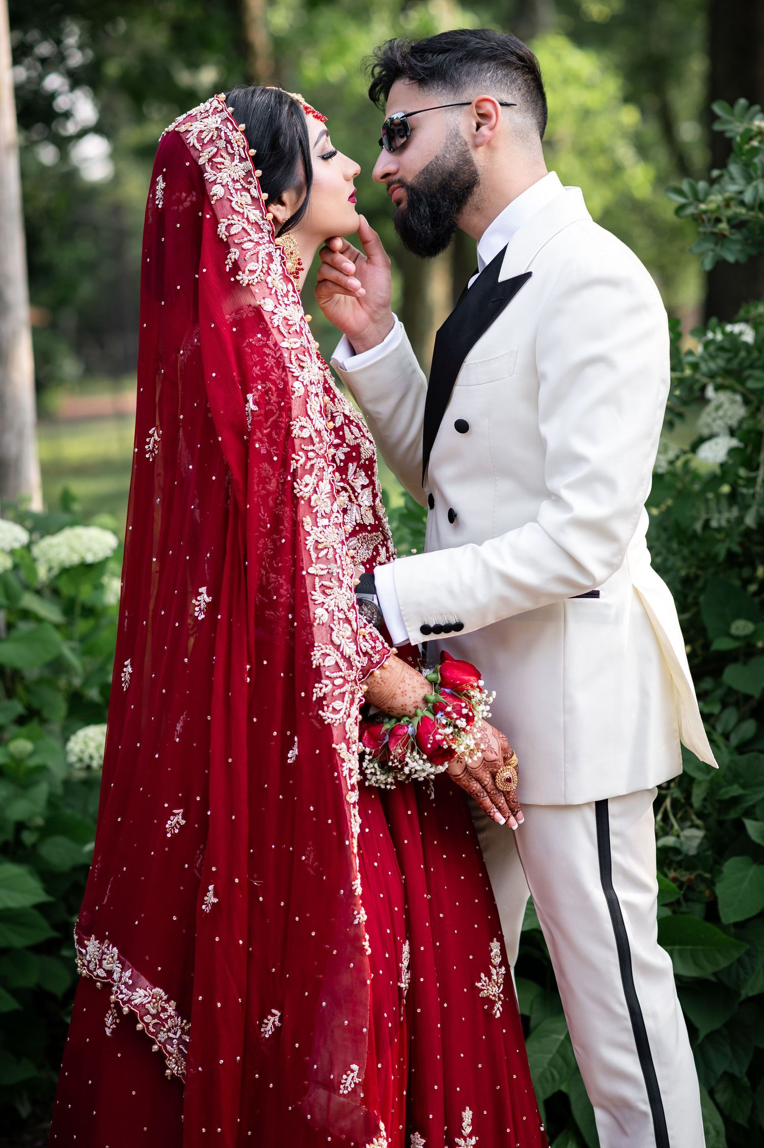Wedding couple in outdoor garden setting with bride in red embroidered dress and groom in white suit sharing intimate moments.