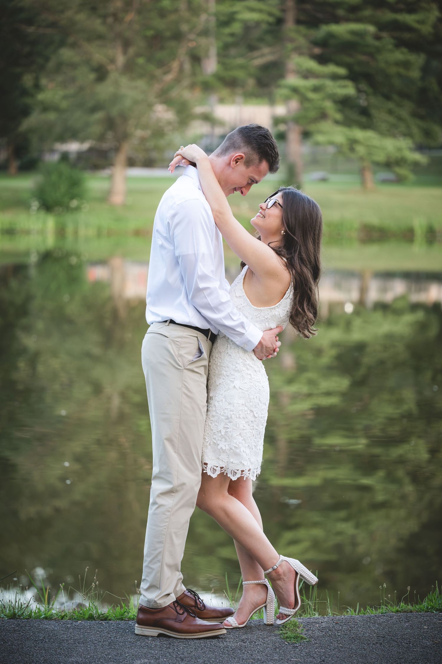 Couple embracing romantically in white lace dress and khakis beside a serene reflective pond in a lush park.
