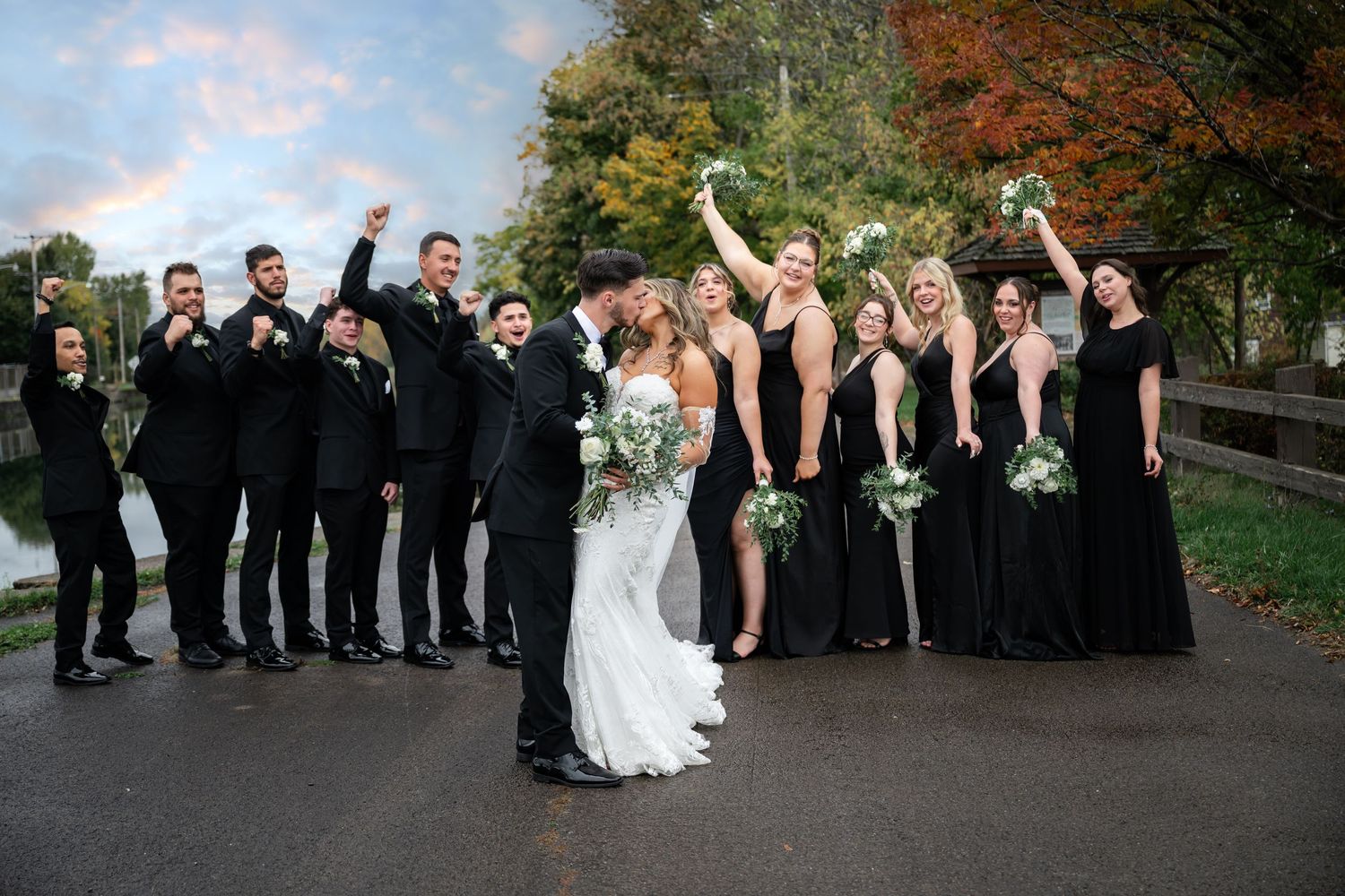 Wedding party in formal attire poses outdoors on a fall day with autumn foliage in the background.