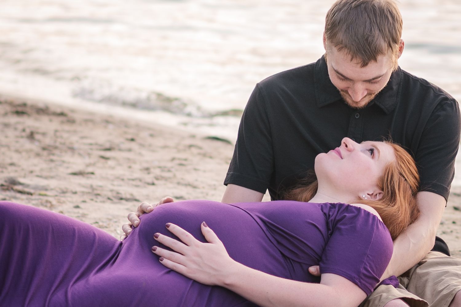 A couple sits together on a sandy beach, with one person wearing a purple dress laying back while the other leans close.