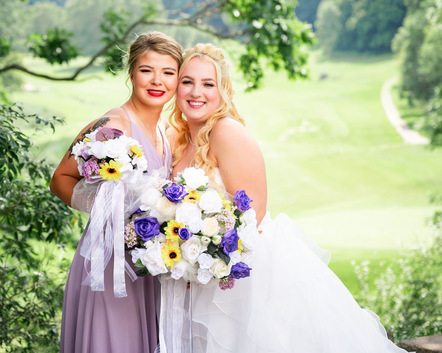 Two wedding party members hold colorful bouquets while posing against a scenic green backdrop.