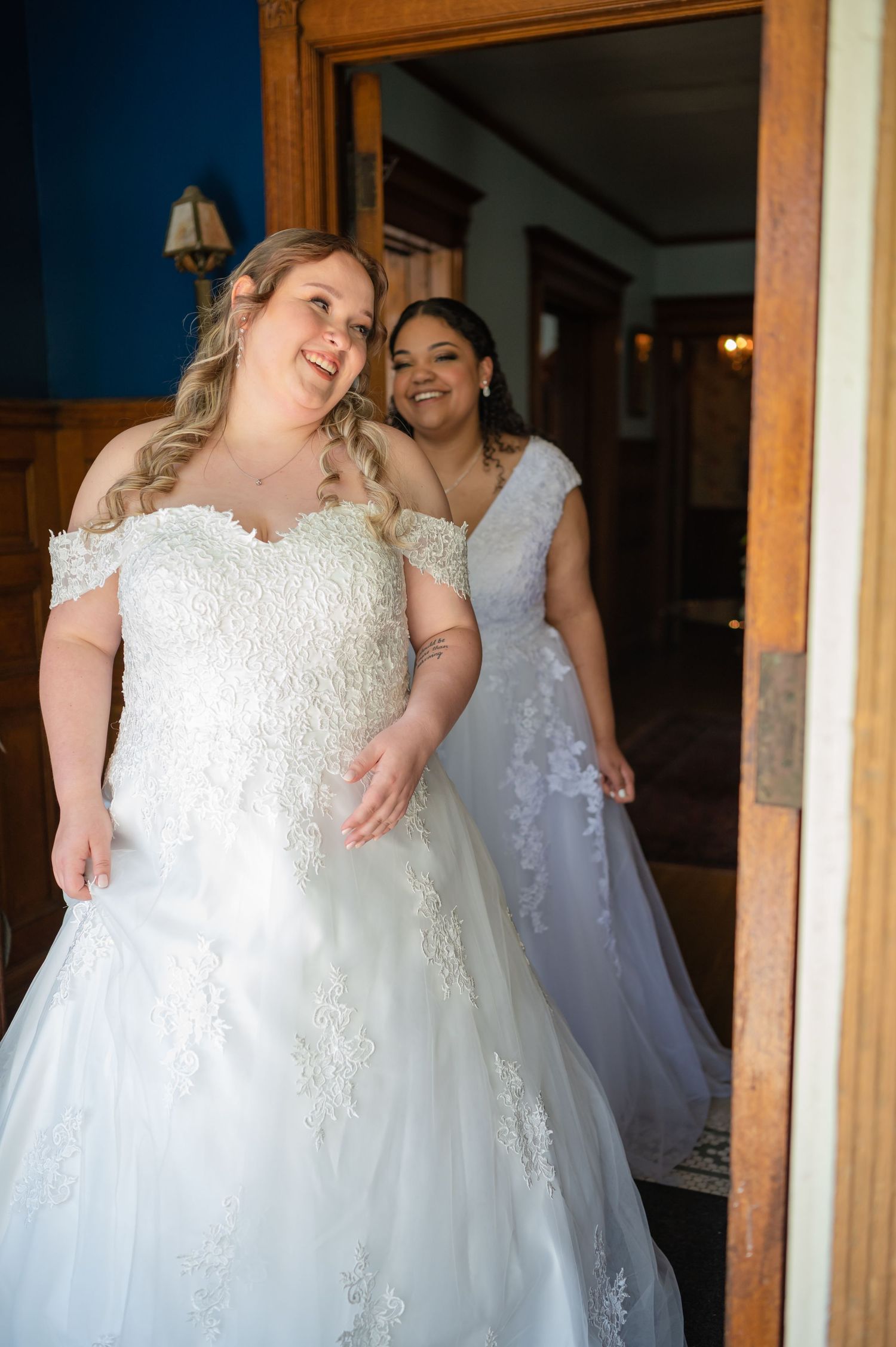 Two people in wedding gowns share a joyful moment in front of a wooden doorway.