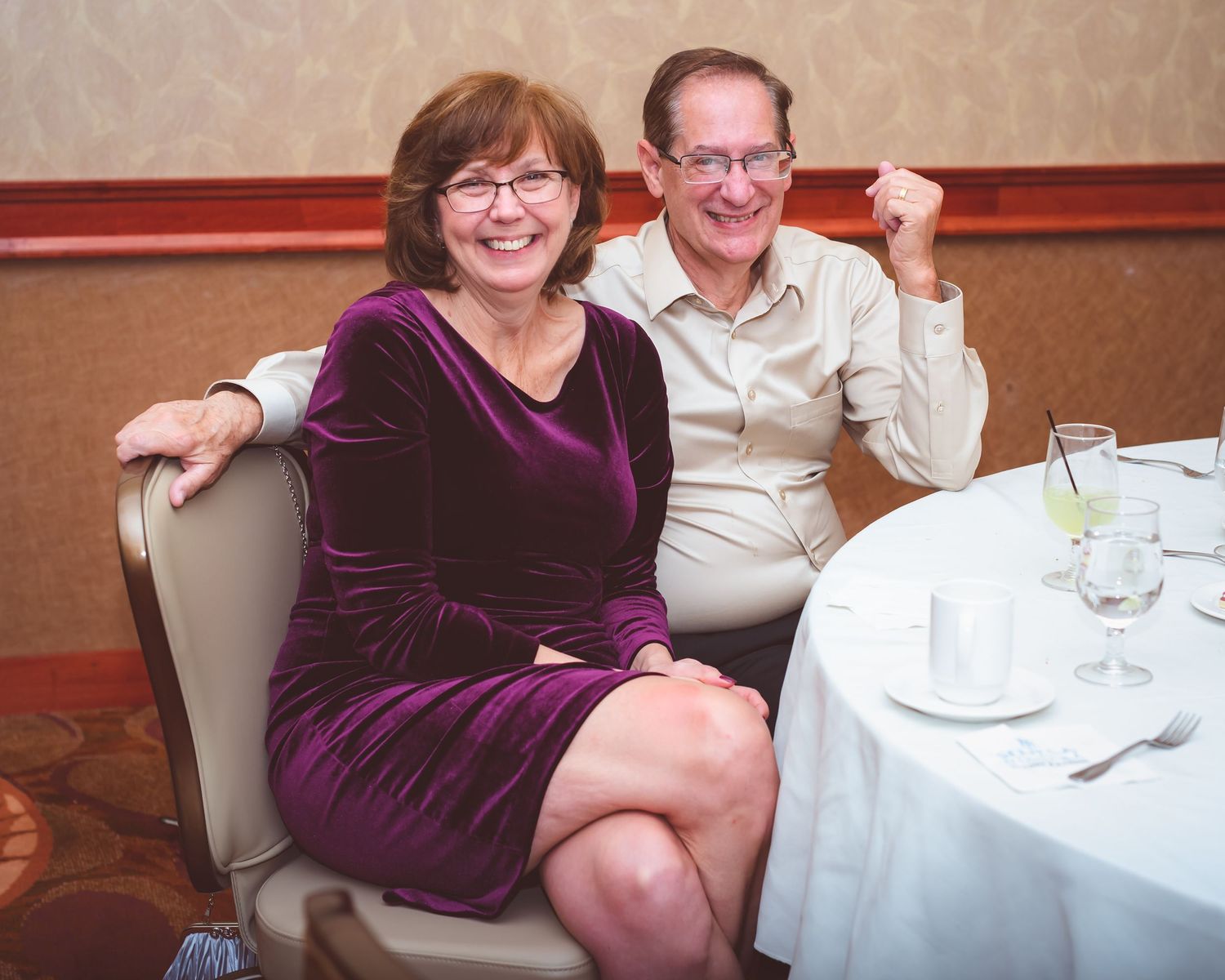 A couple in formal attire smiles while seated at a round dining table.