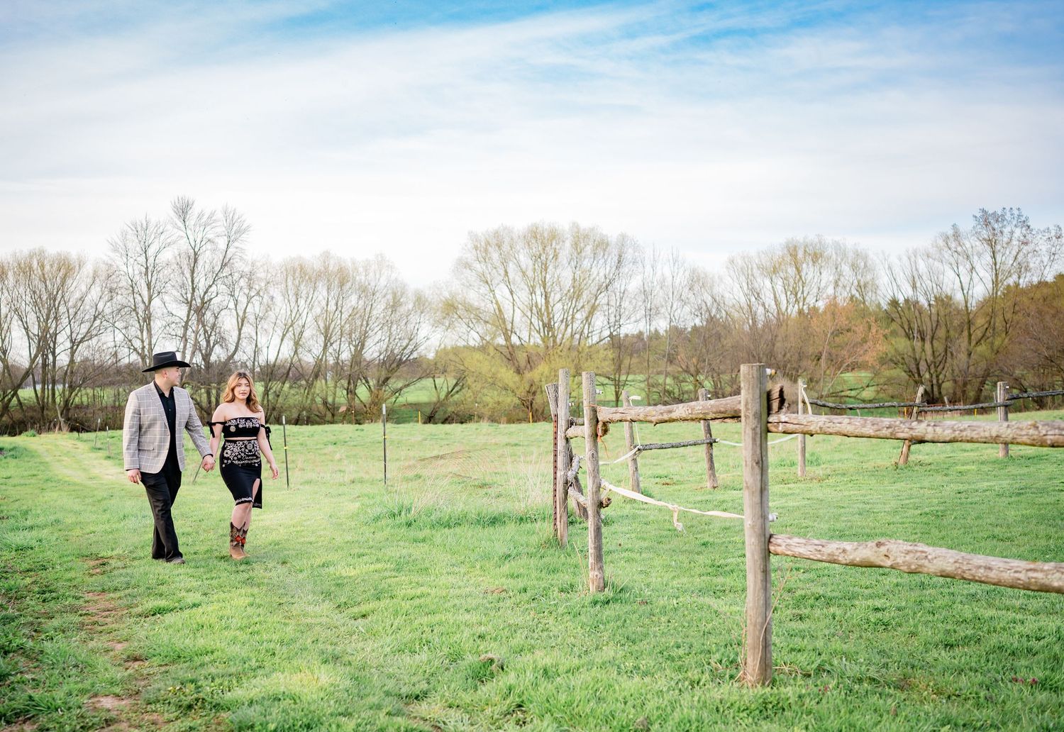 A couple walking through a green pasture with wooden fence posts under a blue sky.
