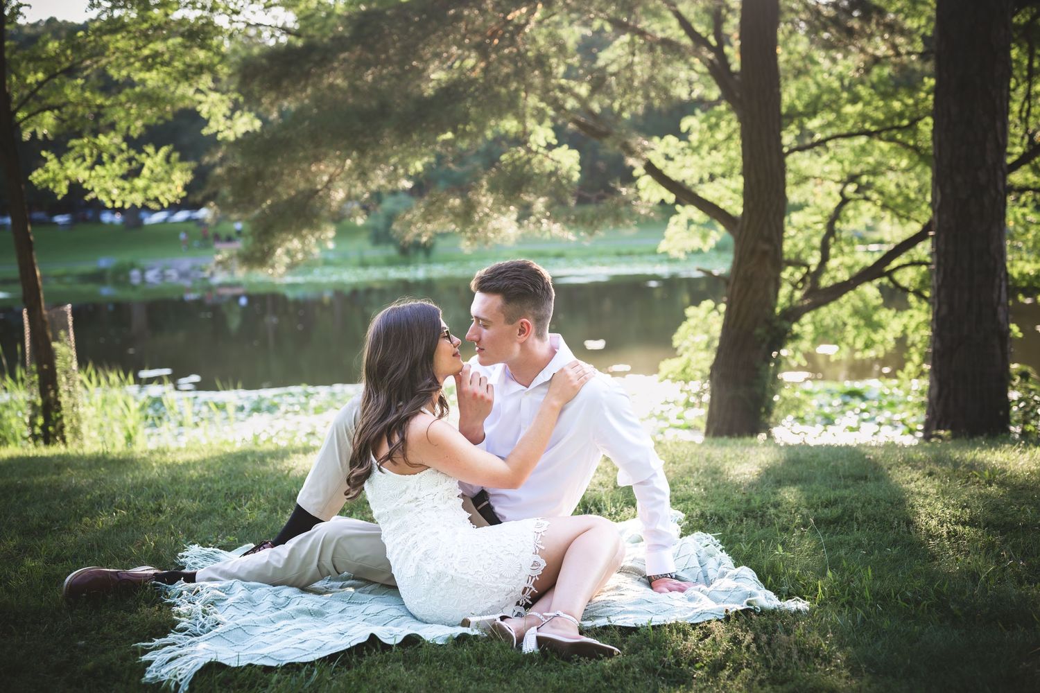 Couple sitting on a blanket by a serene lake in a lush green park, sharing a romantic moment outdoors.