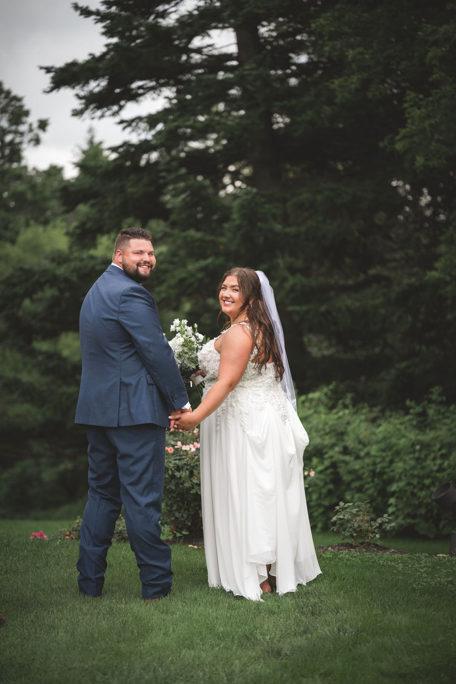 Bride and groom holding hands in lush garden, bride in white lace gown with veil, groom in navy suit.