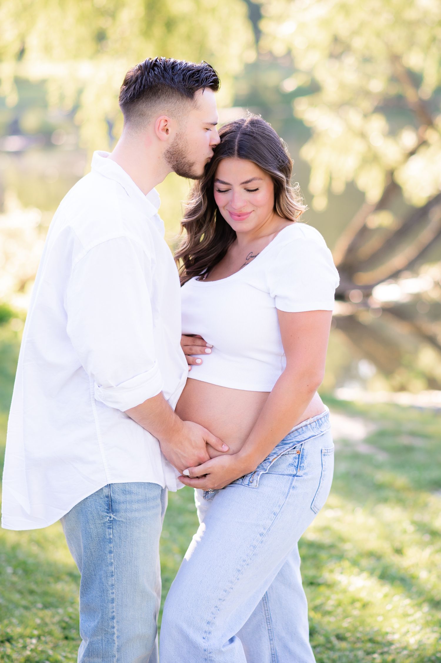 Couple in white tops and jeans sharing a tender moment outdoors, woman showing pregnant belly, man kissing her head.